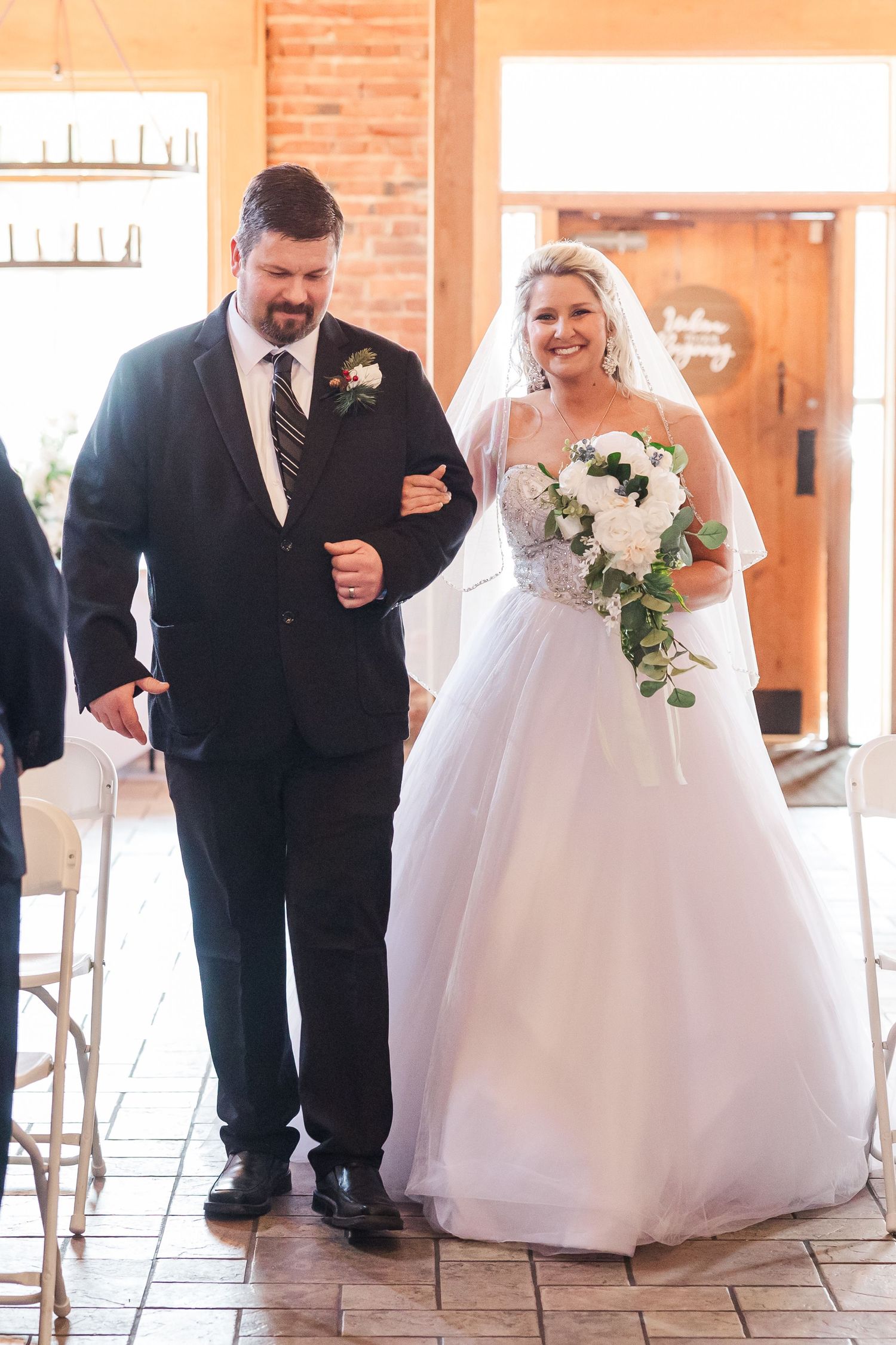 A traditional wedding processional moment in an indoor venue with exposed brick walls and white decorations.
