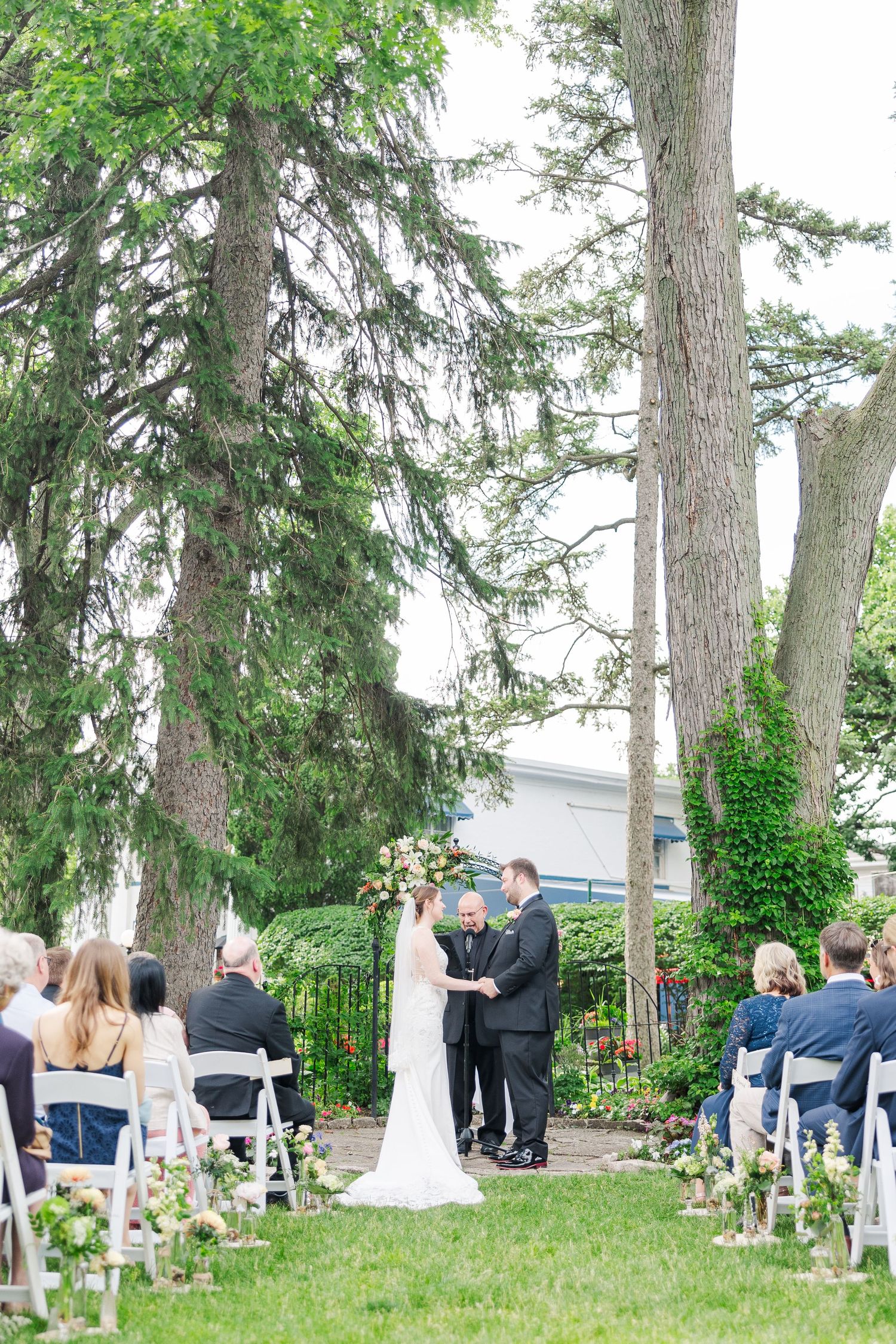 Outdoor wedding ceremony setup with white chairs under tall evergreen trees.