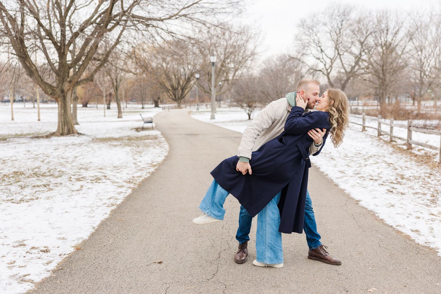 A couple dips playfully on a winding path through a snowy park landscape with bare winter trees. Proposal Winnemac Park, Chicago
