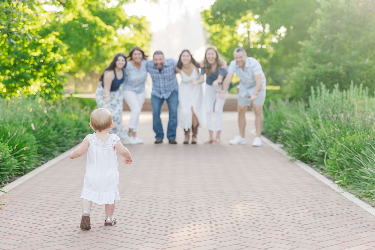 A small child in a white dress runs down a brick path toward a group of people gathered in a garden setting at sunset.