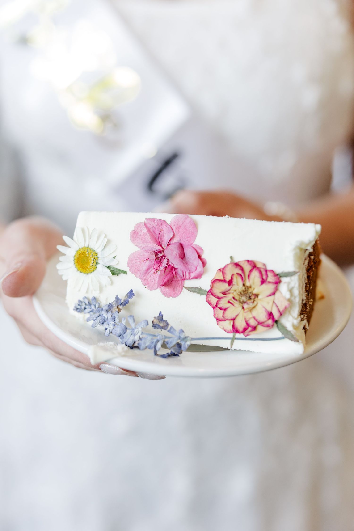 A hand holding a white plate with a slice of cake decorated with pressed pink and white edible flowers.