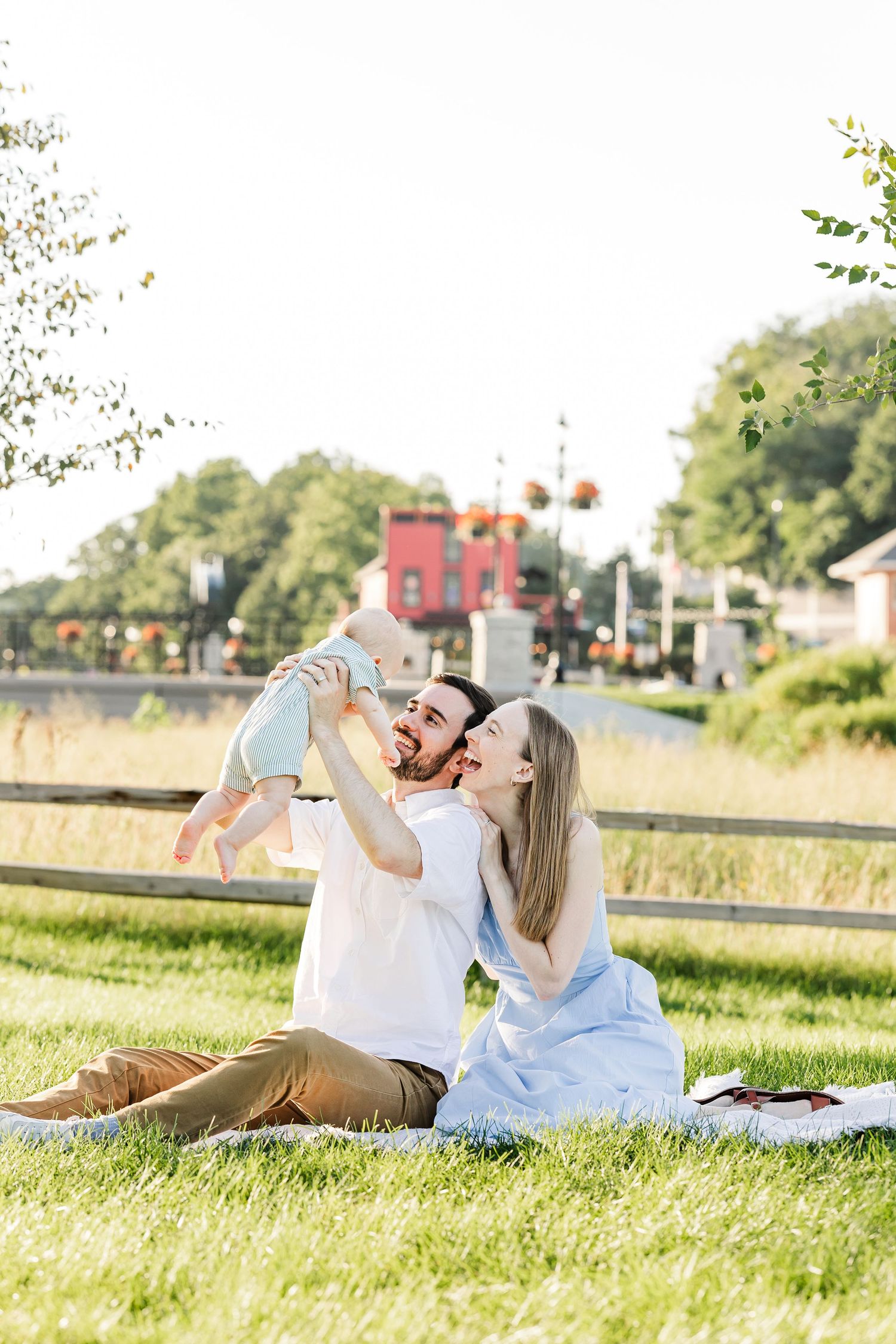 Happy couple sitting on grass in a sunny park, playfully lifting their smiling baby together.