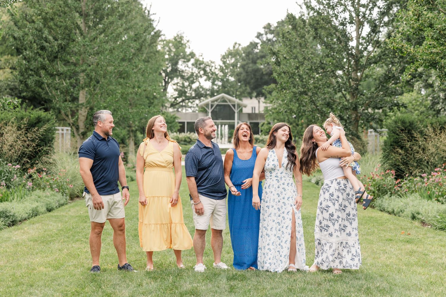 A joyful family of seven laughs together outdoors in a lush garden setting, dressed in colorful summer attire.