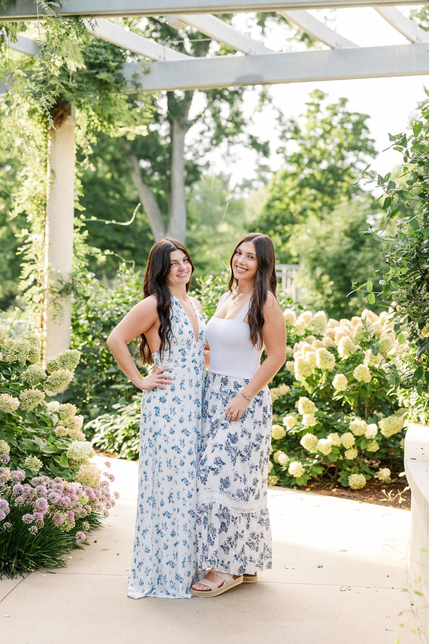 Two young women in floral maxi skirts pose under a pergola surrounded by lush greenery and white hydrangeas.