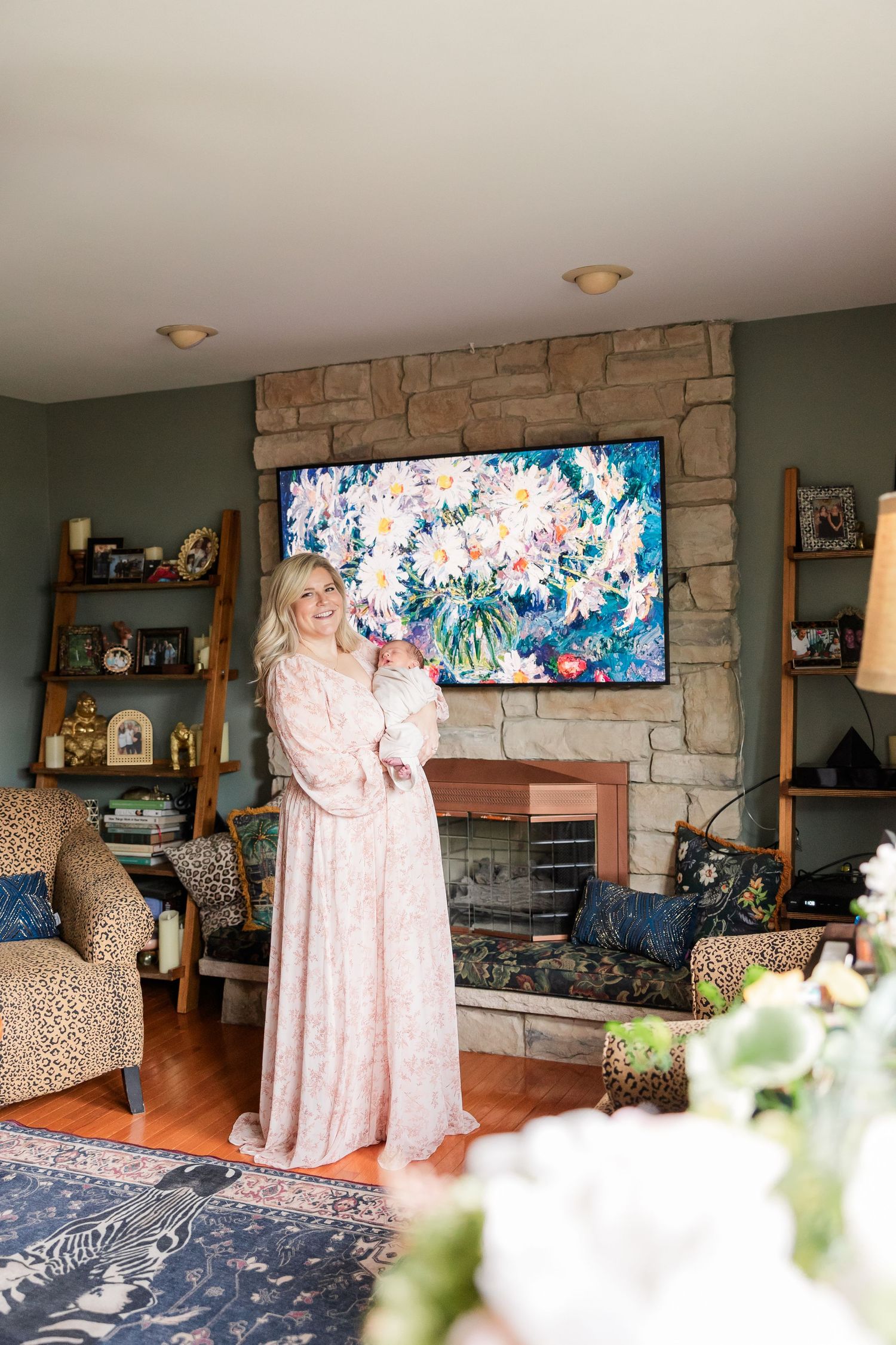 Woman in pink floral gown holding baby by stone fireplace with floral art above it in cozy living room.
