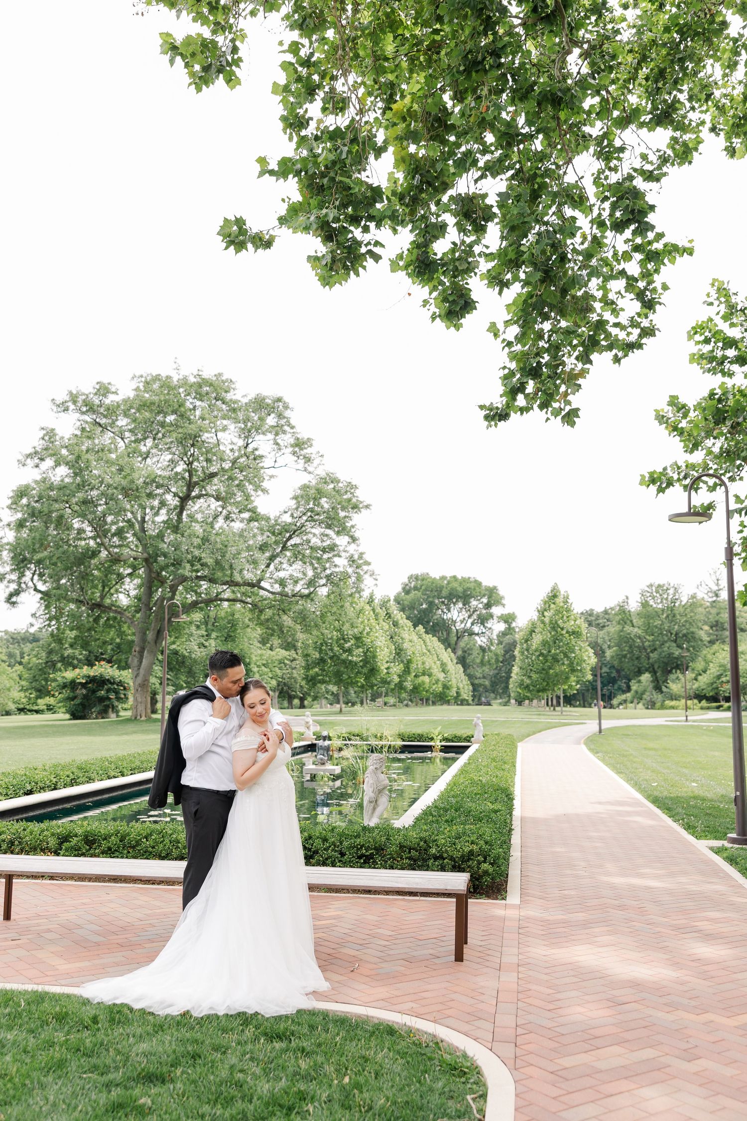Monika and Rafael standing together along a garden path surrounded by trees and formal landscaping at Cantigny Park.