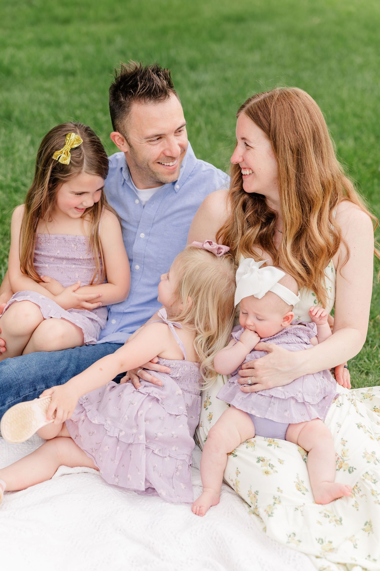 Happy family of five sitting on a blanket in green grass, parents smiling at each other with three young daughters in purple dresses.