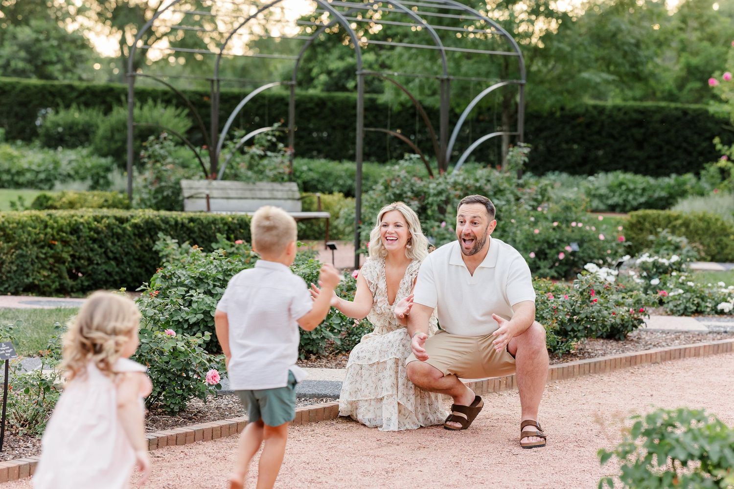 Family with two children laughing and playing together in a rose garden at sunset.