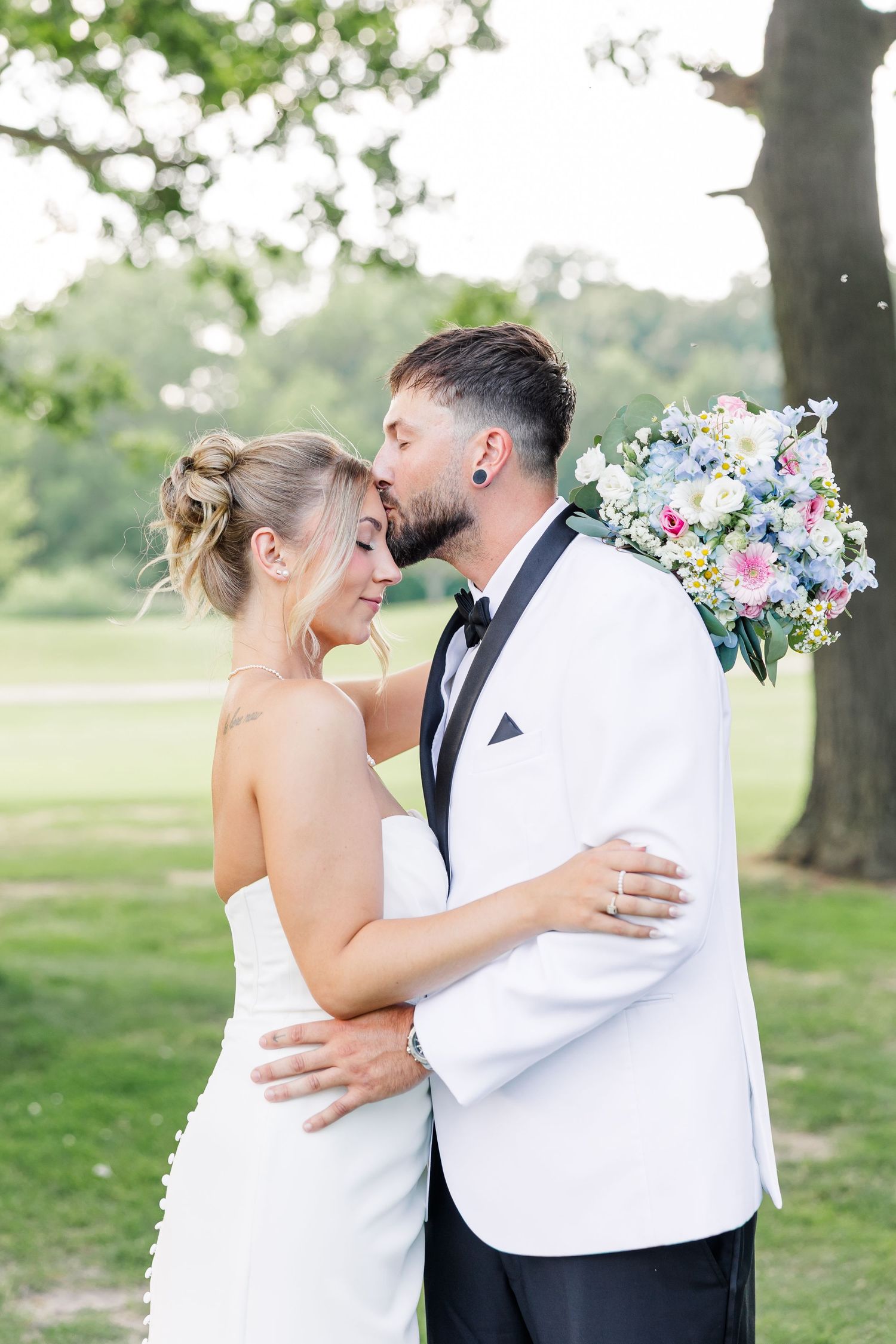 A bride and groom share a tender kiss outdoors, the groom holding a pastel floral bouquet in a white tuxedo.