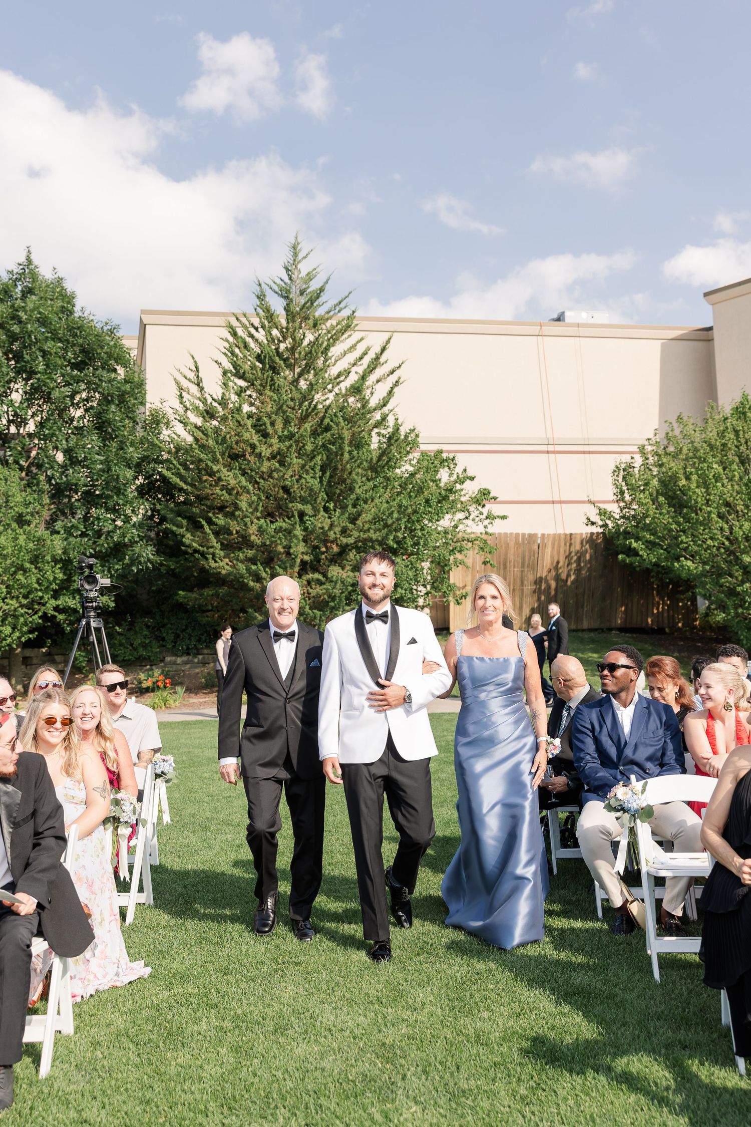 Groom in white tuxedo walks down outdoor aisle escorted by parents during sunny wedding ceremony.