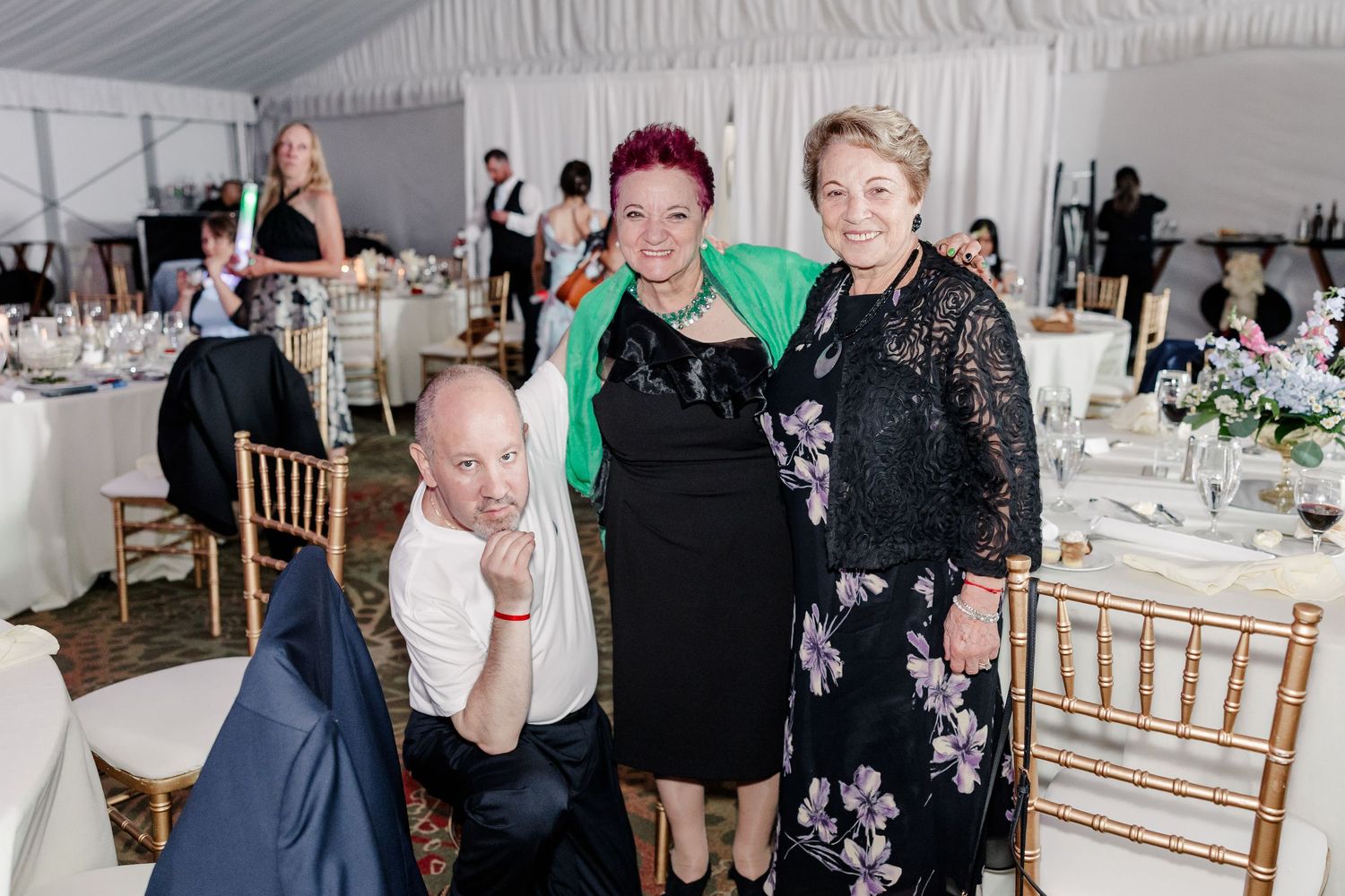 Three wedding guests pose and smile together inside an elegant white tent wedding reception venue.