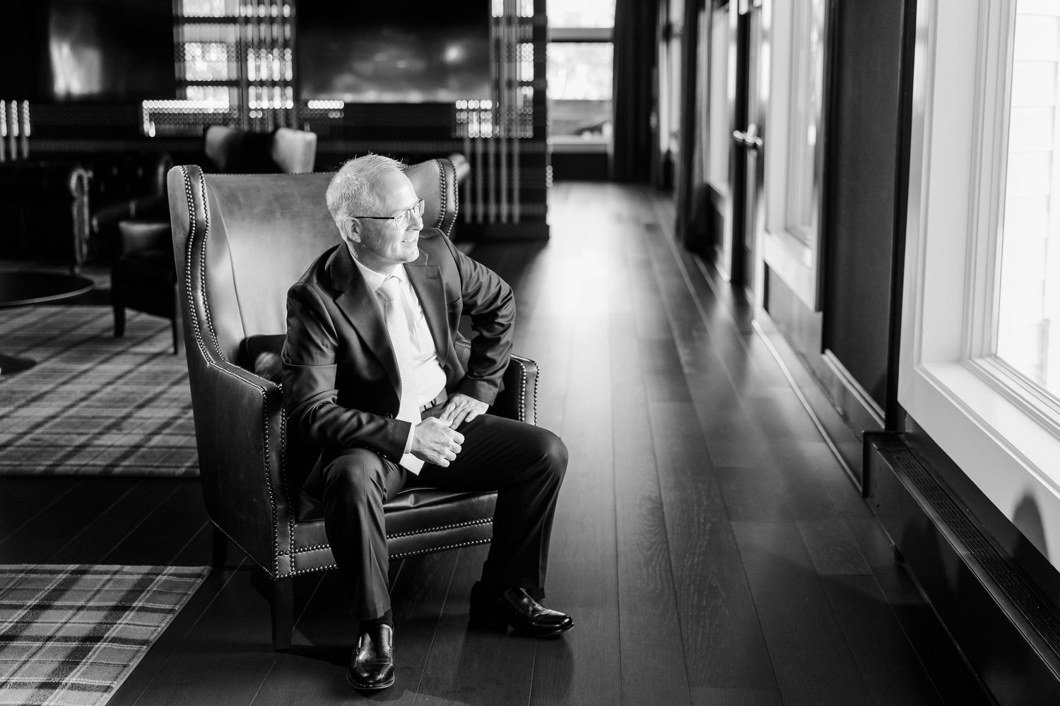 Black and white portrait of a silver-haired man in a suit seated in a leather armchair beside tall windows in an elegant hardwood-floored ro
