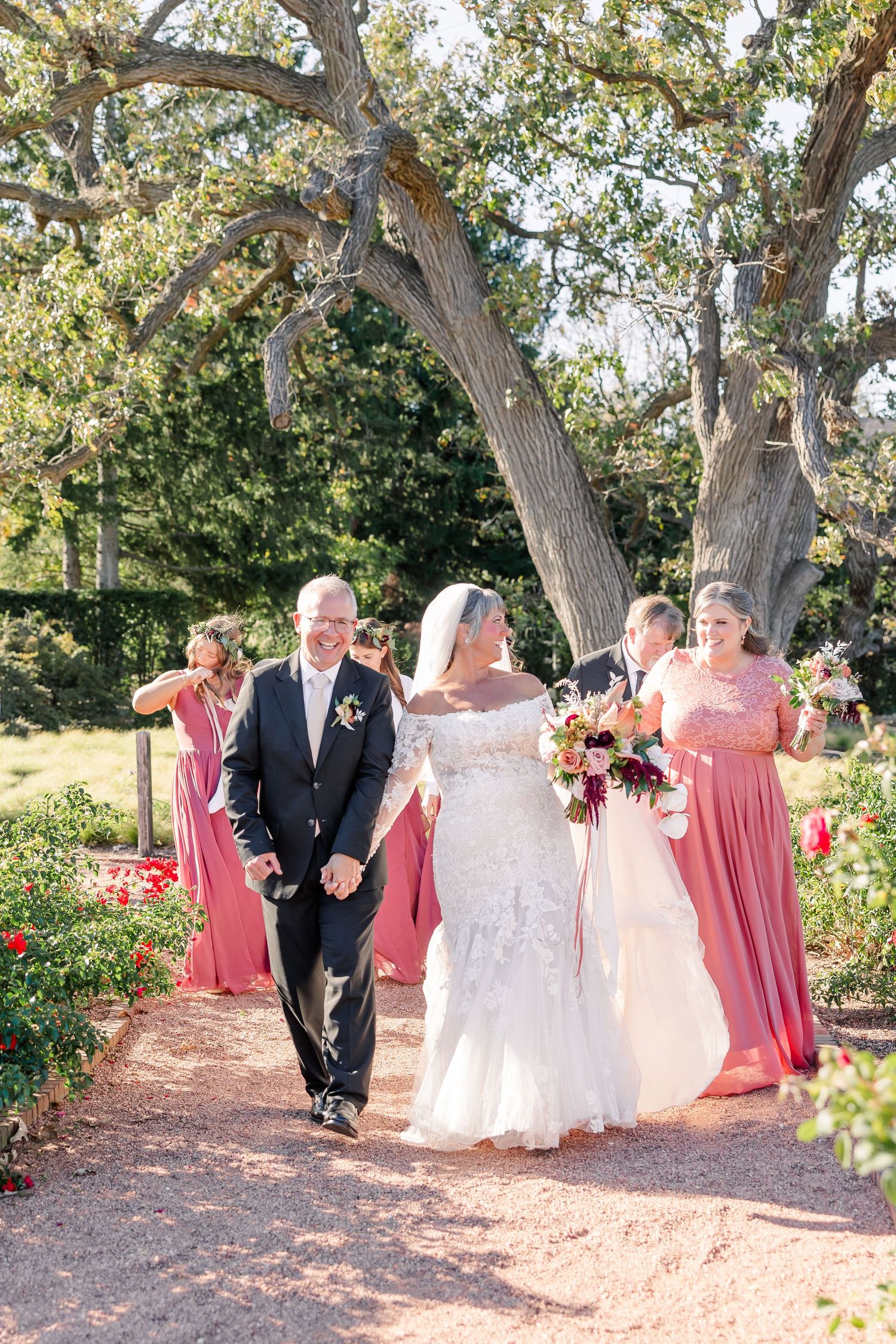 A bride in a lace wedding gown and a groom in a dark suit walk arm in arm along a garden path beneath oak trees with bridesmaids.
