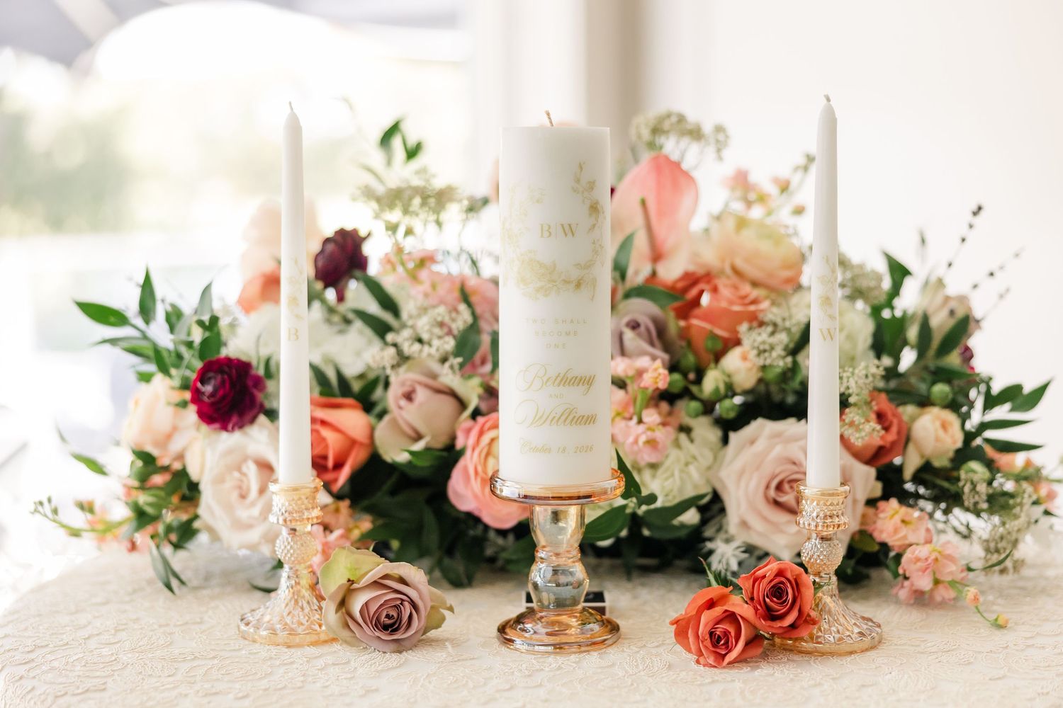 Wedding unity candle with two taper candles on a lace-covered table surrounded by peach and blush roses and greenery.