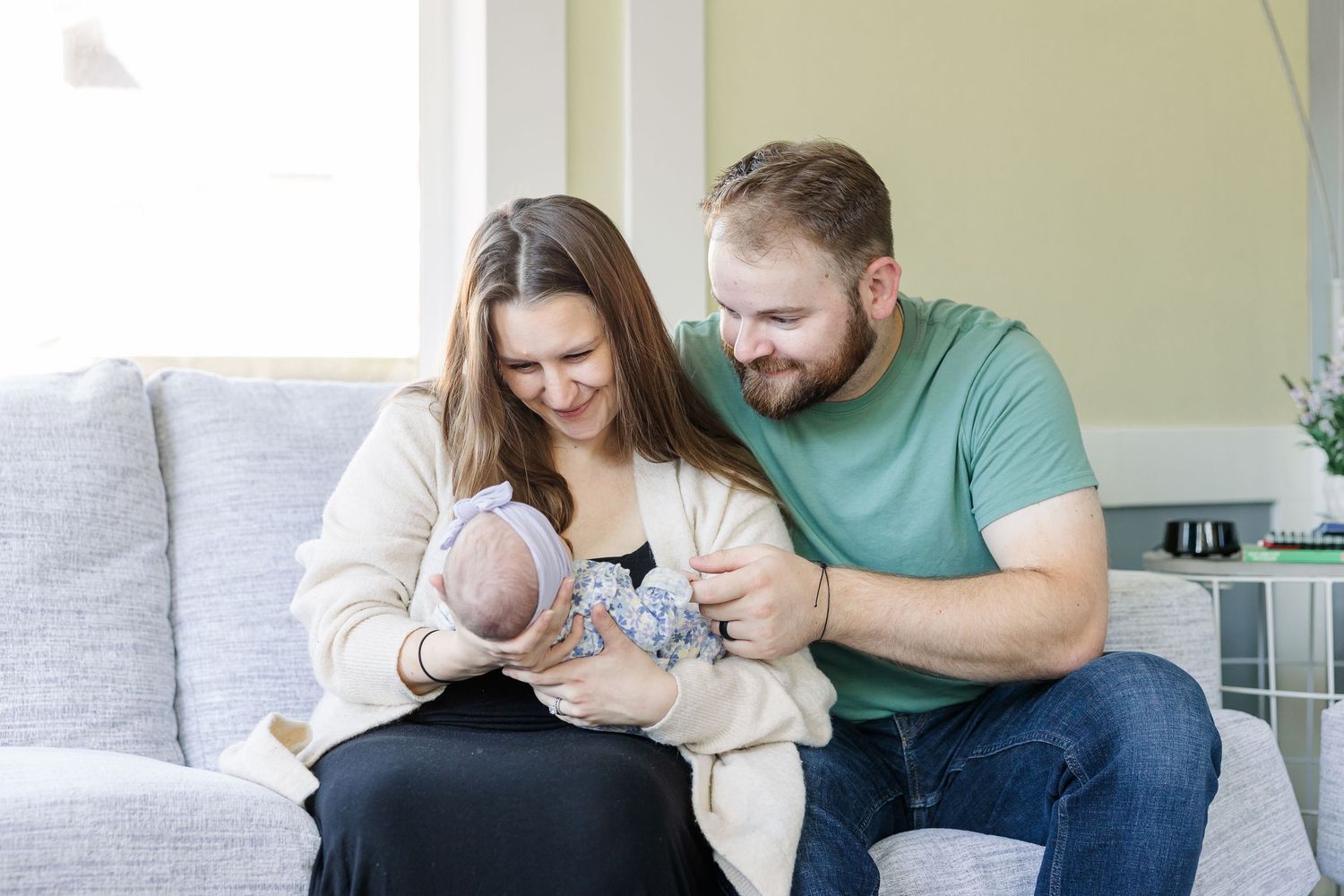 A couple sits on a couch smiling at their newborn baby, with soft natural light in a cozy living room.