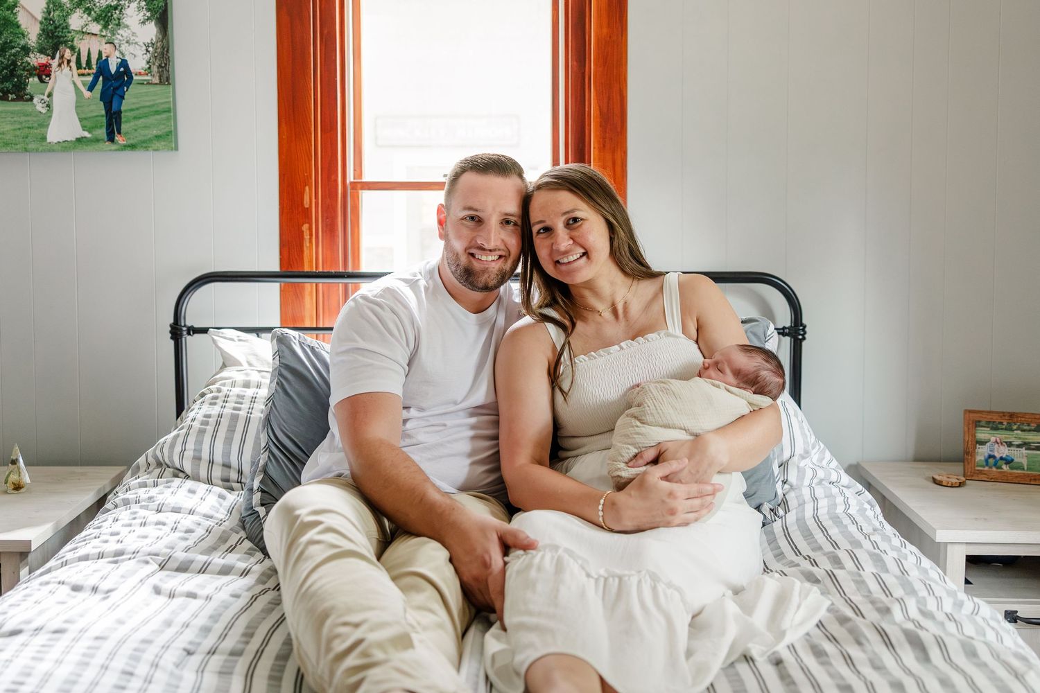 Happy couple sitting on a bed smiling and holding their newborn baby in a cozy bedroom.