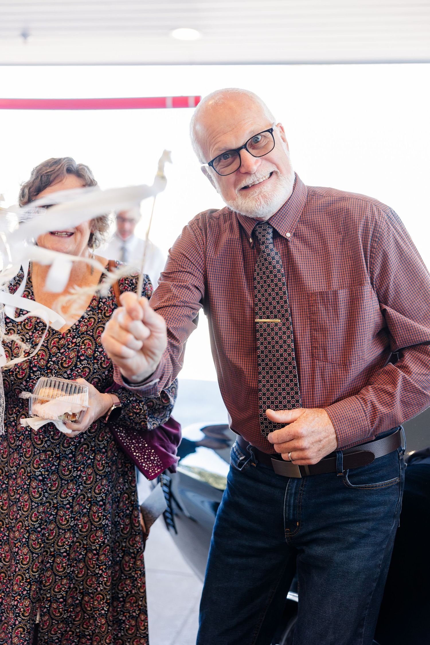 A smiling bearded man in glasses and a maroon shirt holds a ribbon streamer beside a woman in a floral dress at a ribbon-cutting event.