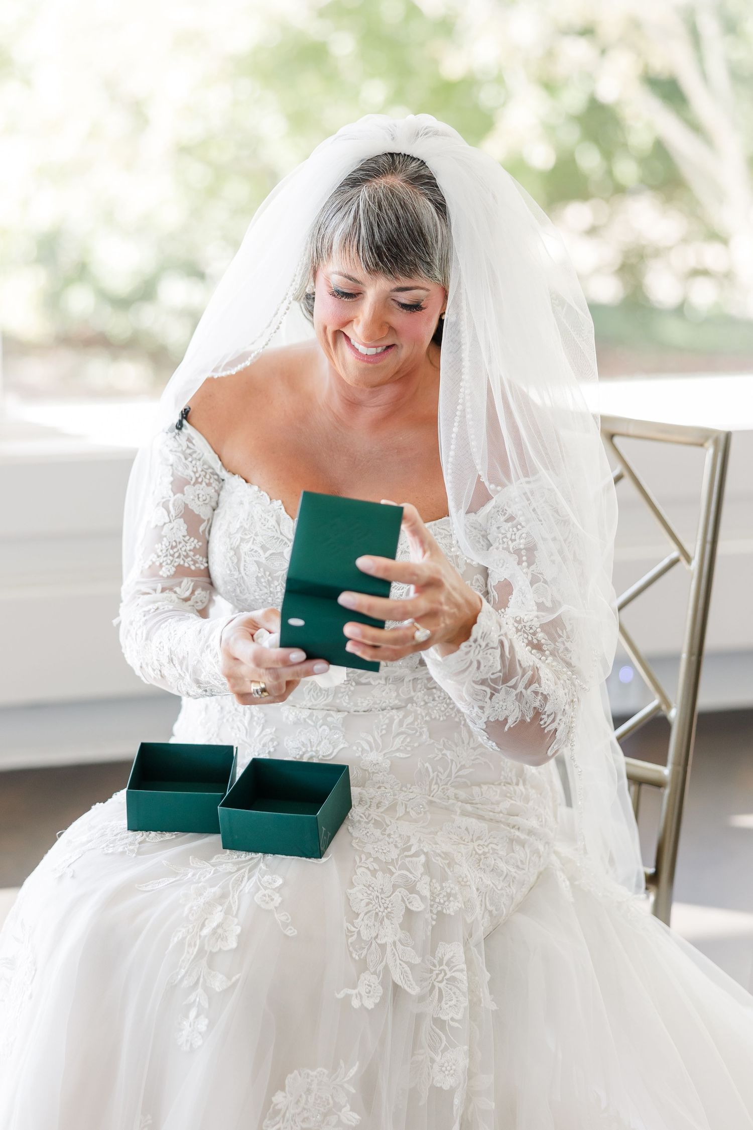 Smiling bride in a lace wedding gown and veil opens a green jewelry box while seated beside a window on her wedding day.