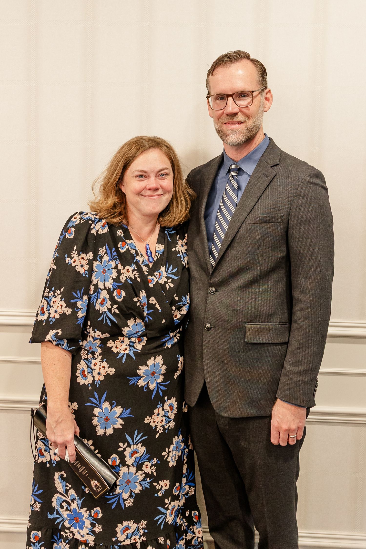 Smiling woman in a navy floral dress stands beside a bearded man in glasses, gray blazer and striped tie at a formal indoor event.