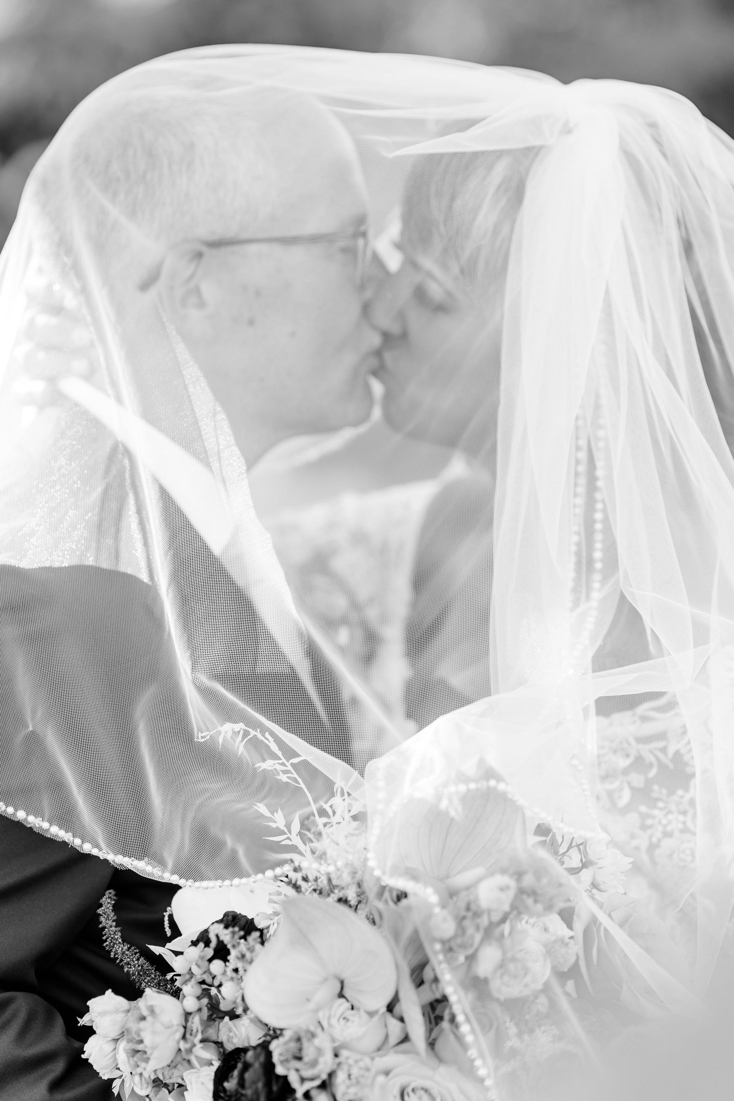 Black and white photo of a couple kissing beneath a sheer wedding veil with a bridal bouquet of flowers in the foreground.