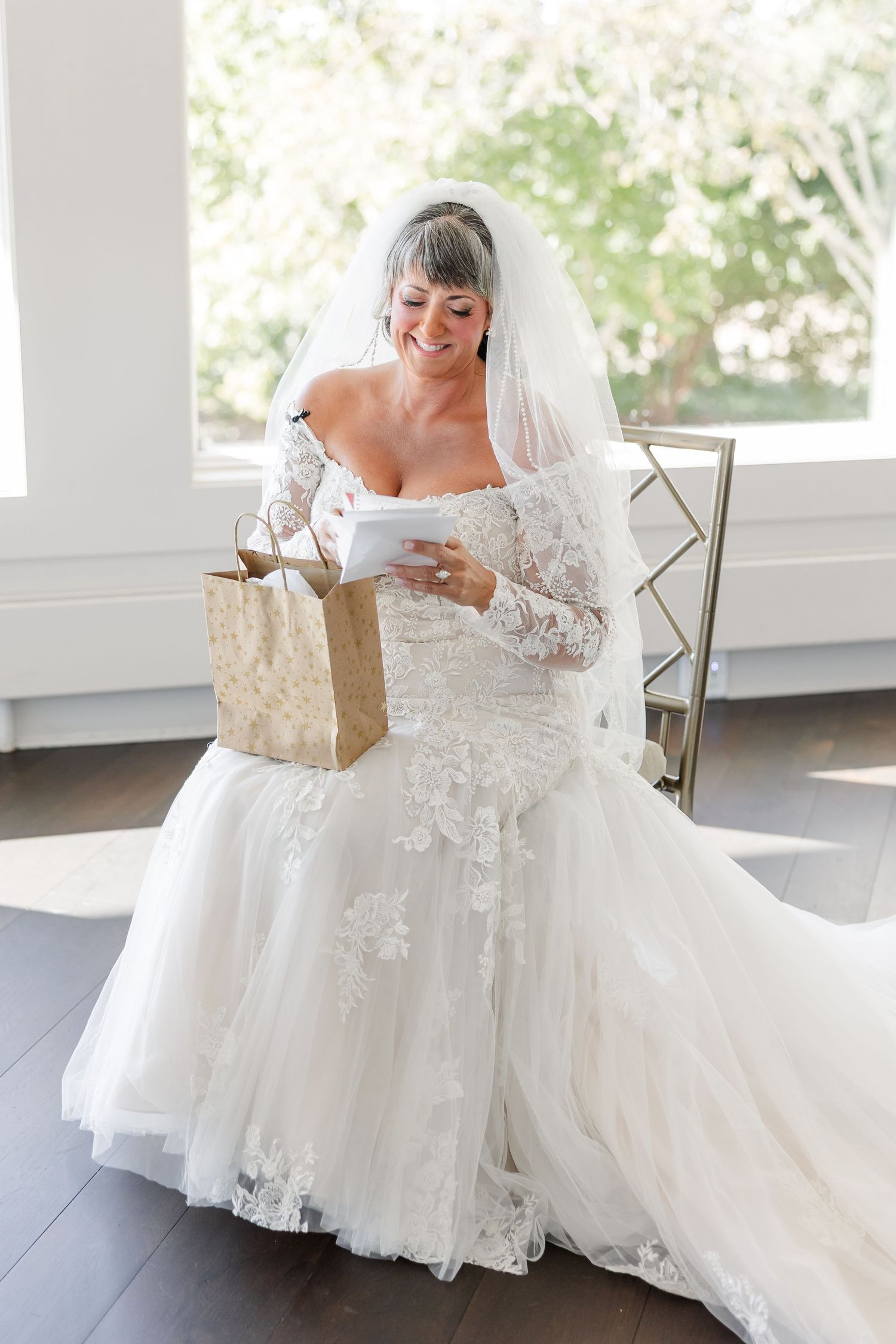 Happy bride in a lace wedding dress and veil sits near a window reading a card beside a gift bag.