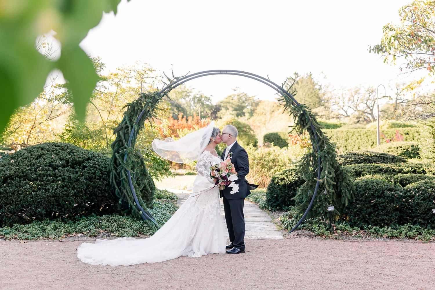 A bride in a white gown and veil kisses her groom beneath a greenery-covered circular arch in a landscaped garden wedding ceremony.