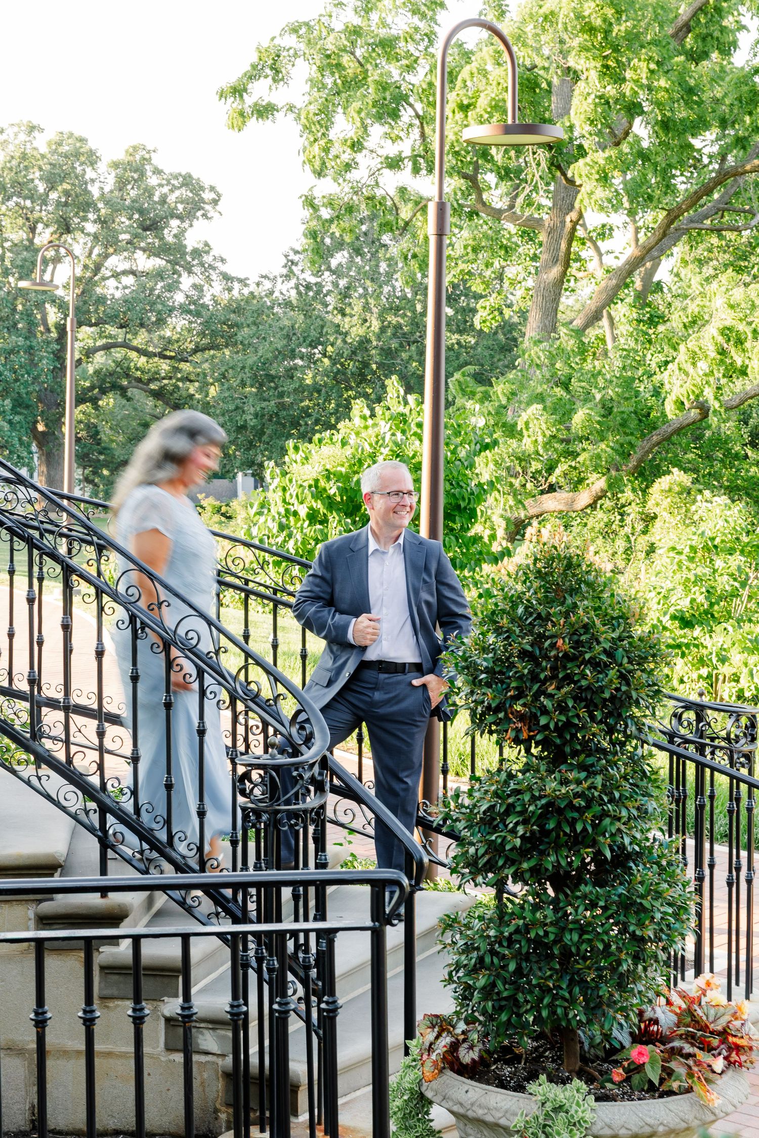 Man in suit smiles at woman on ornate black iron staircase surrounded by lush green garden trees.