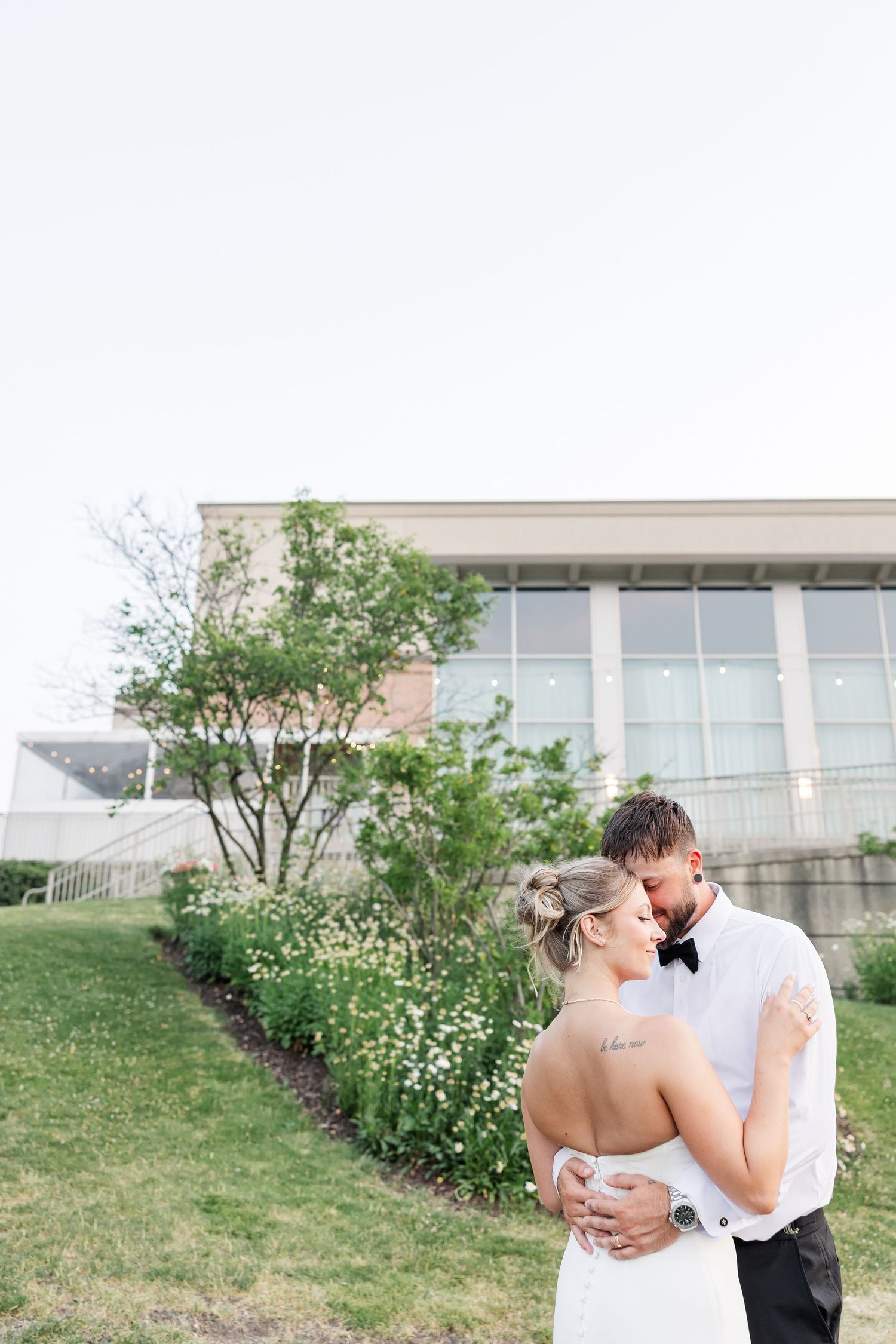 Bride and groom embrace tenderly in front of modern glass building at dusk during wedding.