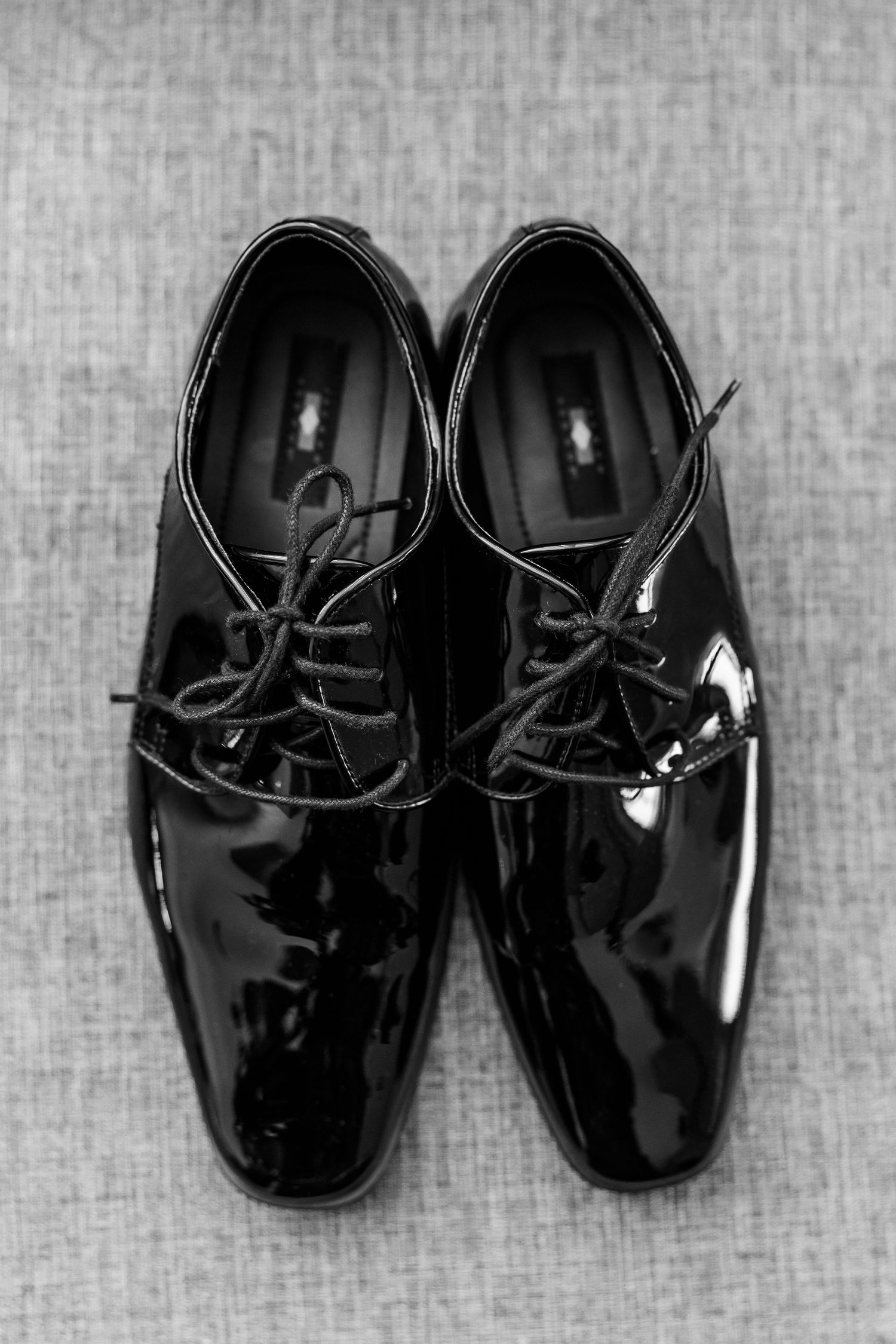 Shiny black patent leather dress shoes with laces on gray fabric background.