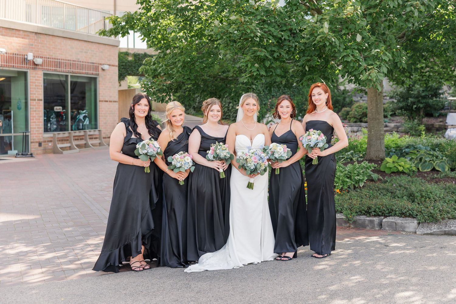 Bride with four bridesmaids in black gowns holding white bouquets posing outdoors.