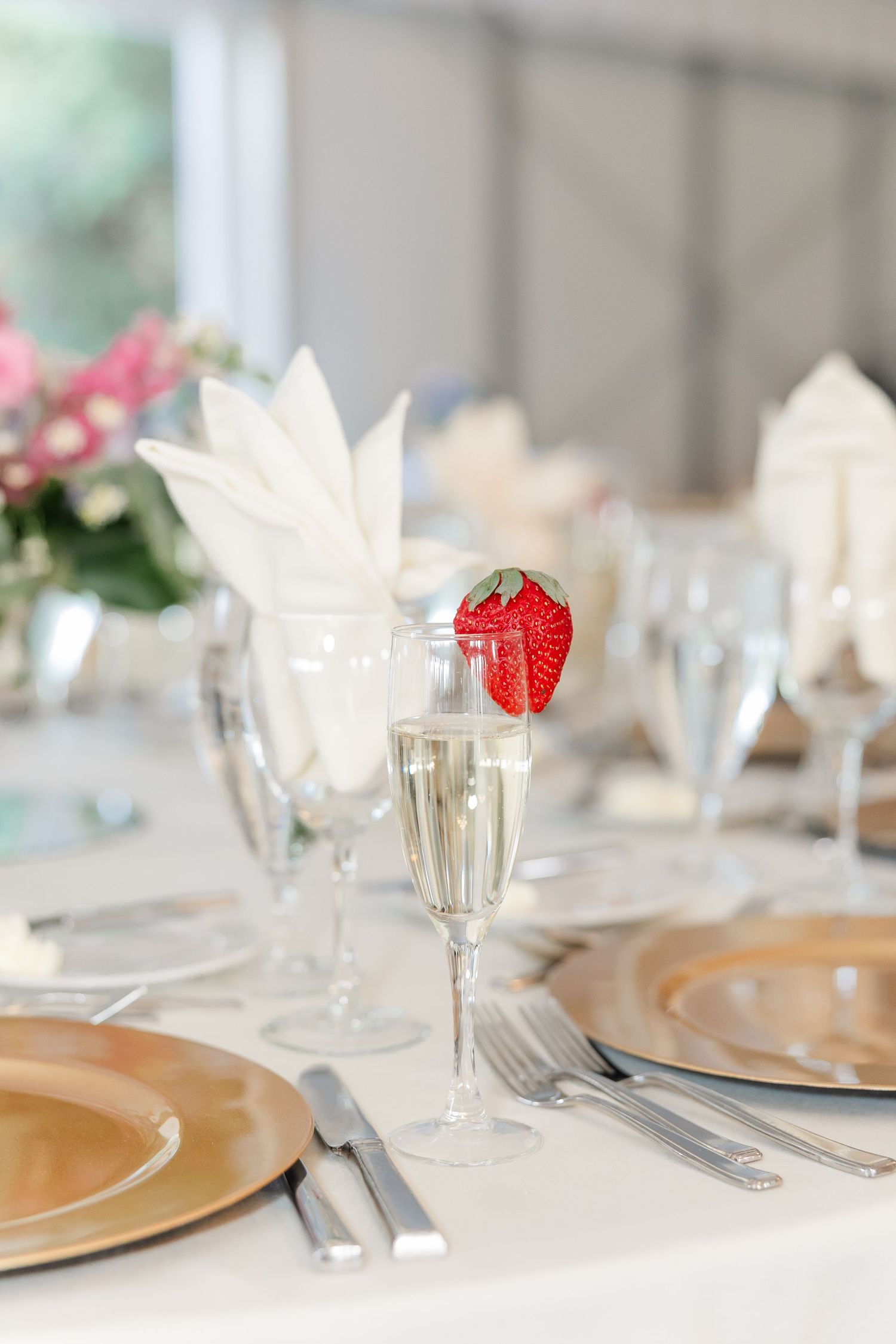 Elegant wedding reception table setting with champagne flute, strawberry garnish, gold charger plates and white napkins.