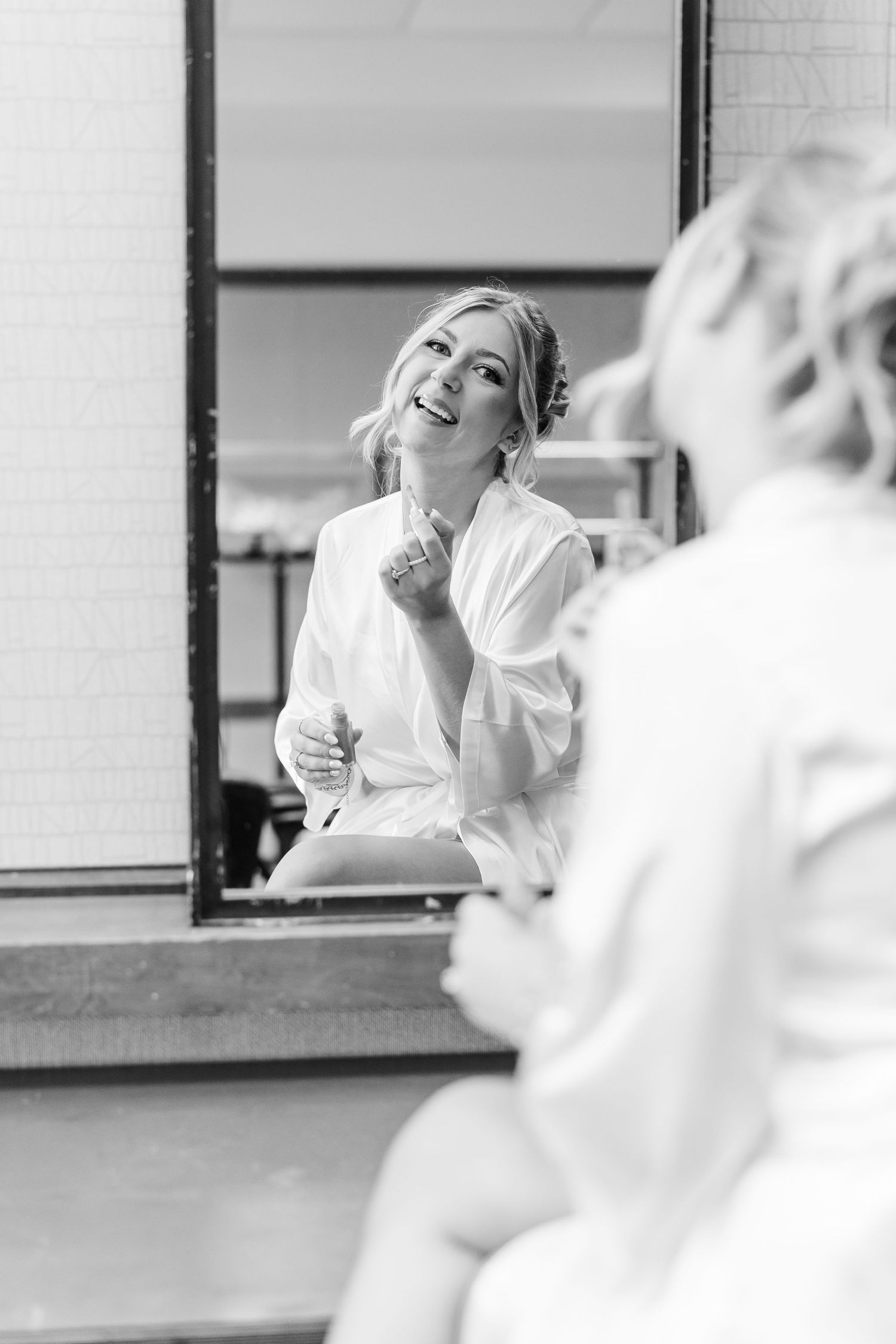 Bride in white robe smiling at her reflection in mirror during wedding morning preparations.