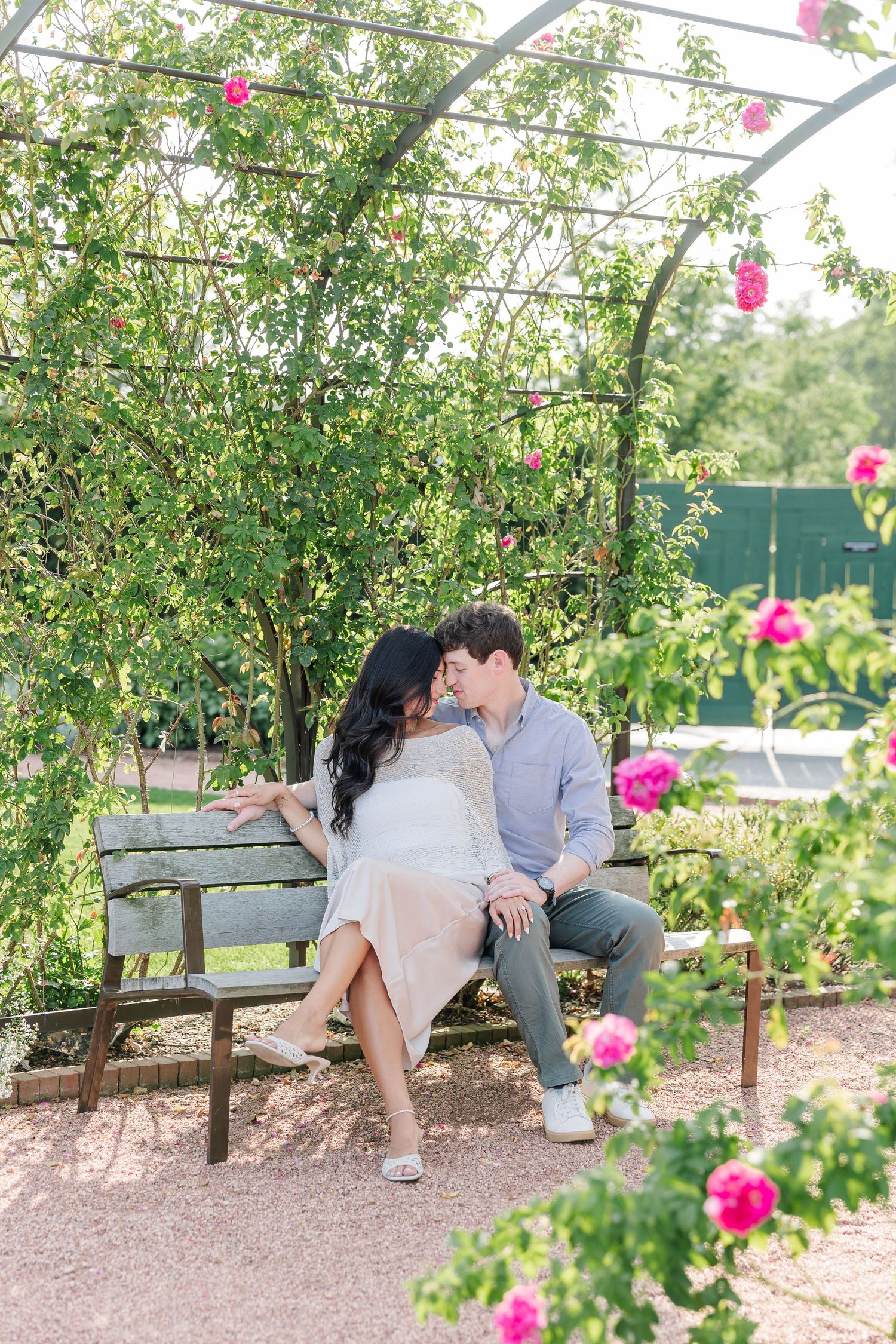 Alysia and Jack share a kiss while sitting on a garden bench surrounded by roses.
