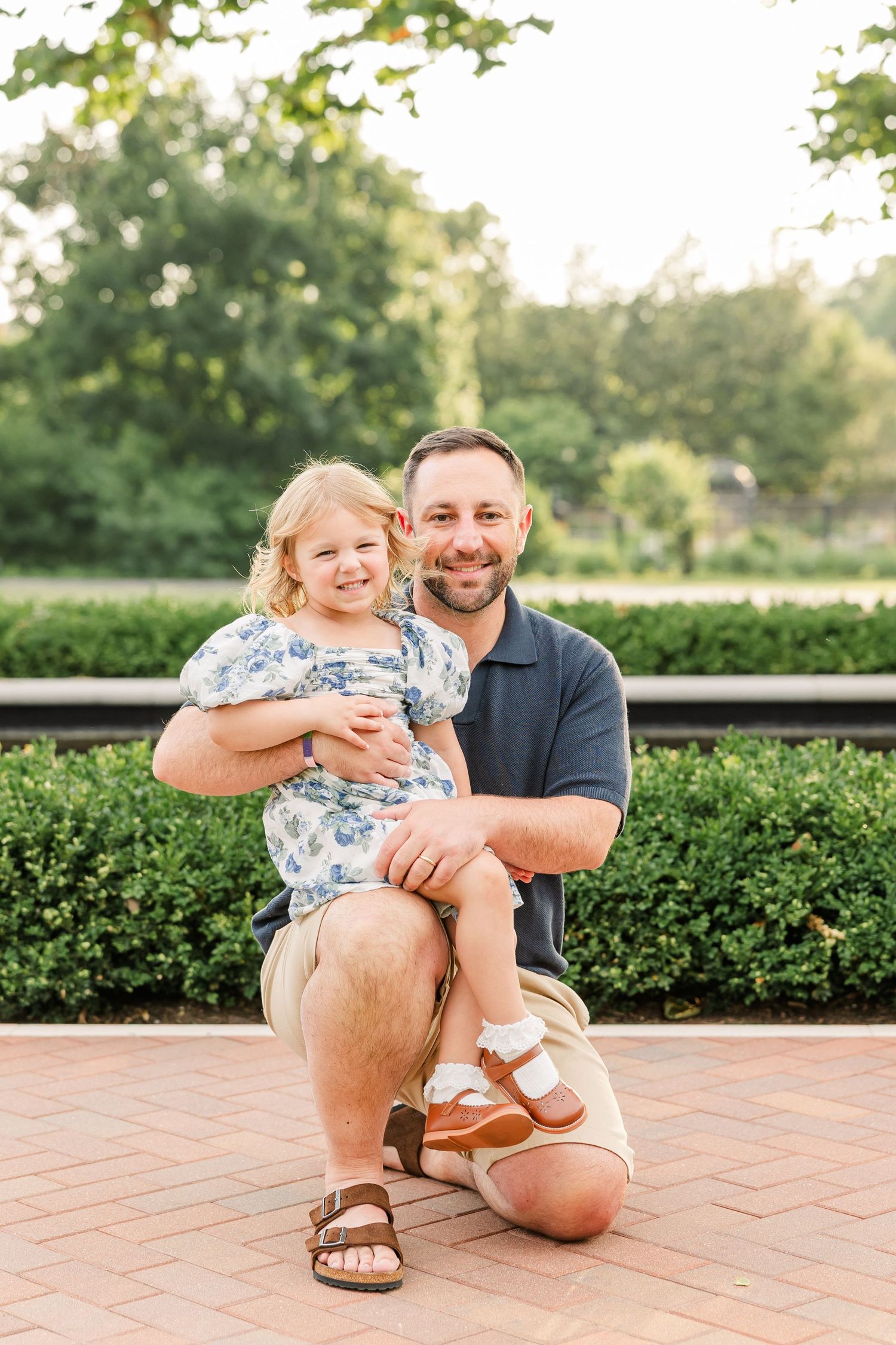 A smiling man crouches on a brick path holding a young blonde girl in a floral dress, surrounded by lush green hedges.