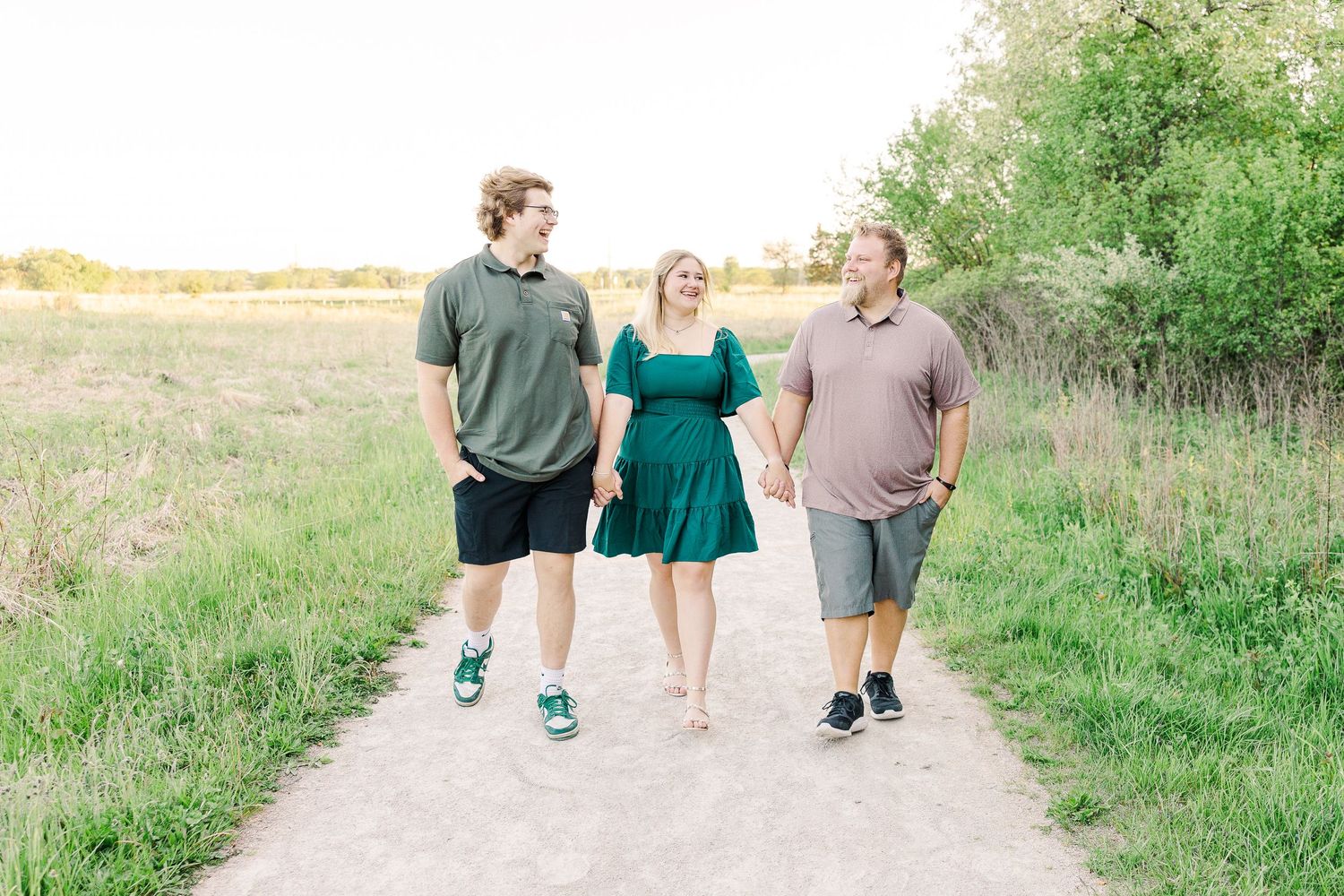 Three people walking together and smiling on a scenic dirt path through grassy fields.