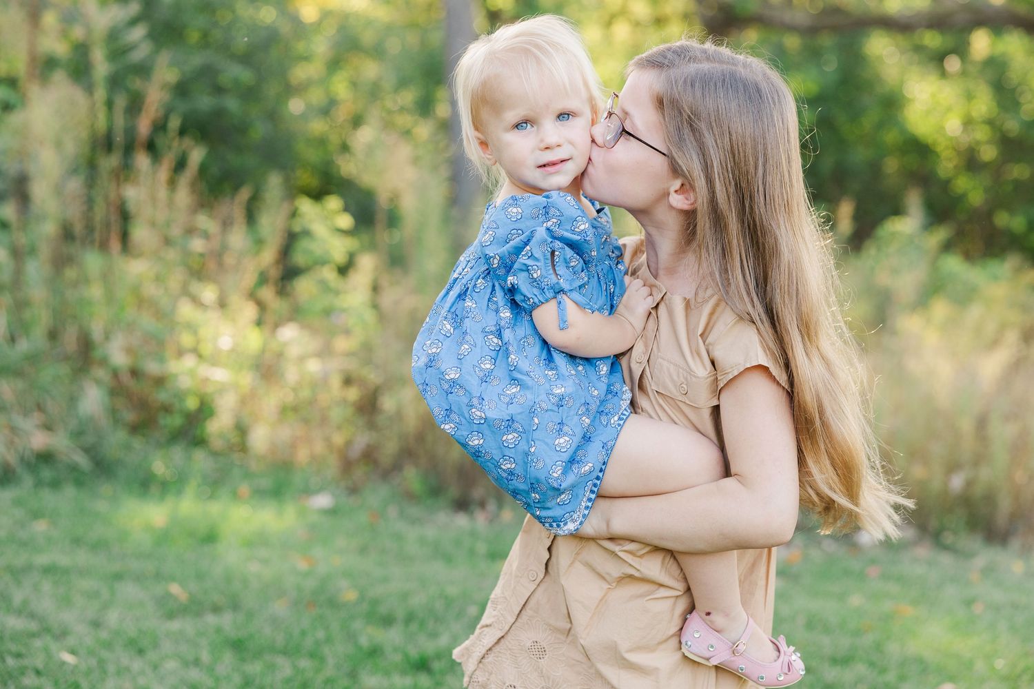 Mother and child in coordinating blue outfits embrace while standing in a sunlit meadow during golden hour.