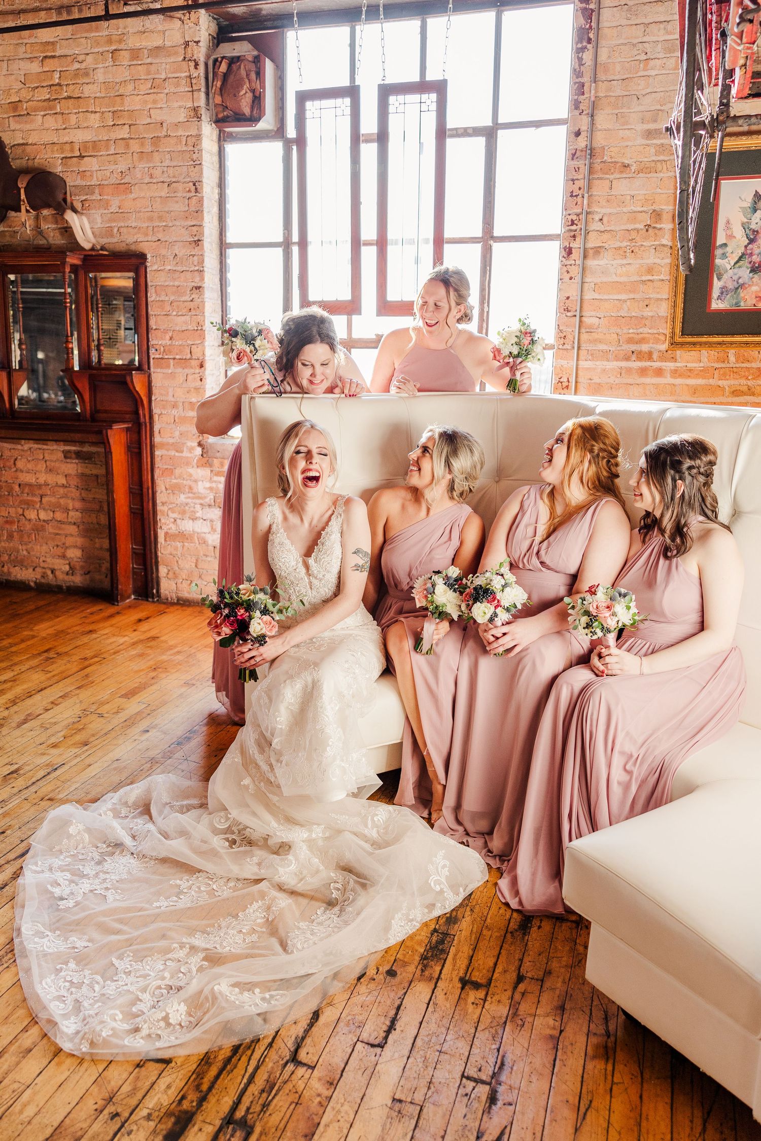 Bridal party shares joyful moment on white couch in rustic brick venue.