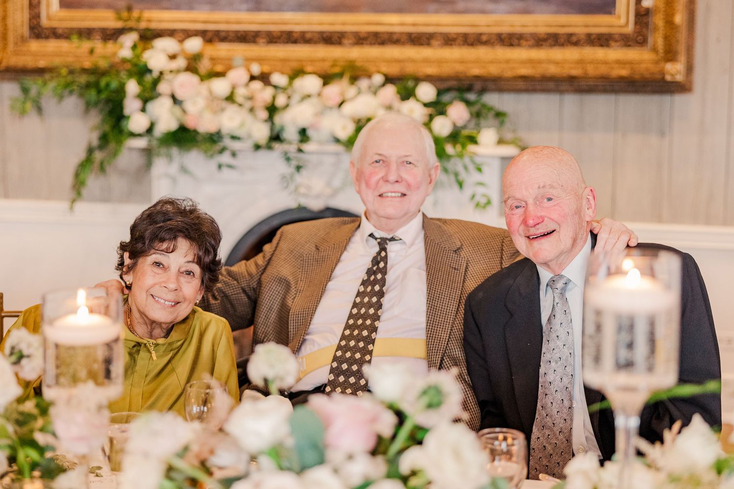 Three guests share a joyful moment at an elegant wedding reception table decorated with floral arrangements.