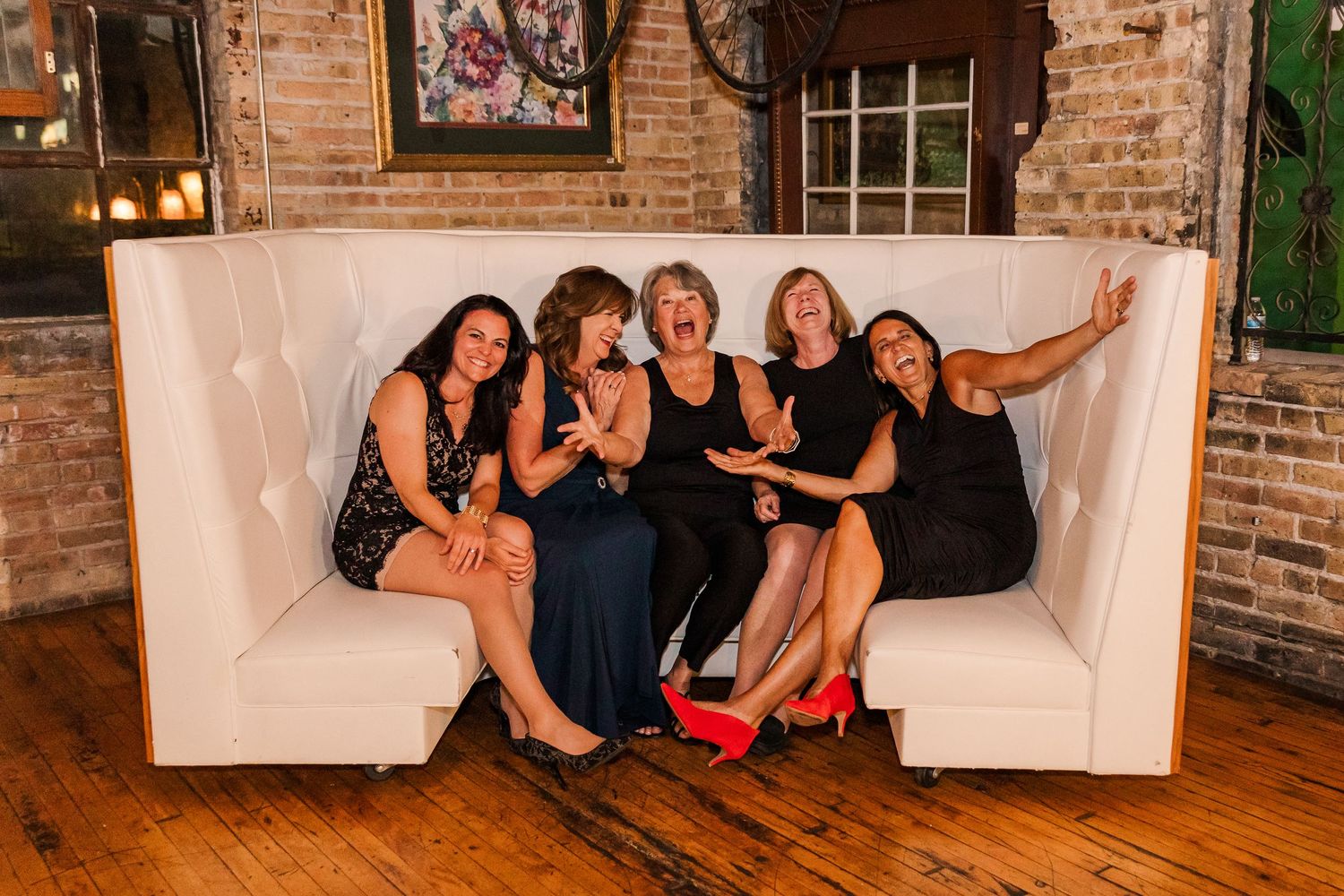 Five women in black dresses strike a fun pose on a white couch at a wedding reception.