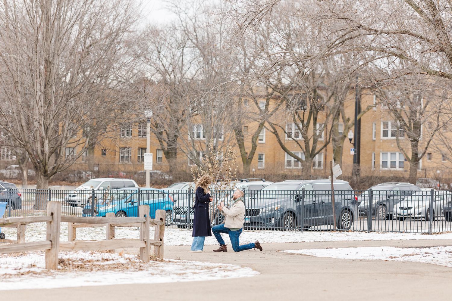 Man proposes to woman in snow-dusted park with bare trees and parked cars visible behind an iron fence in winter. Winnemac Park, Chicago