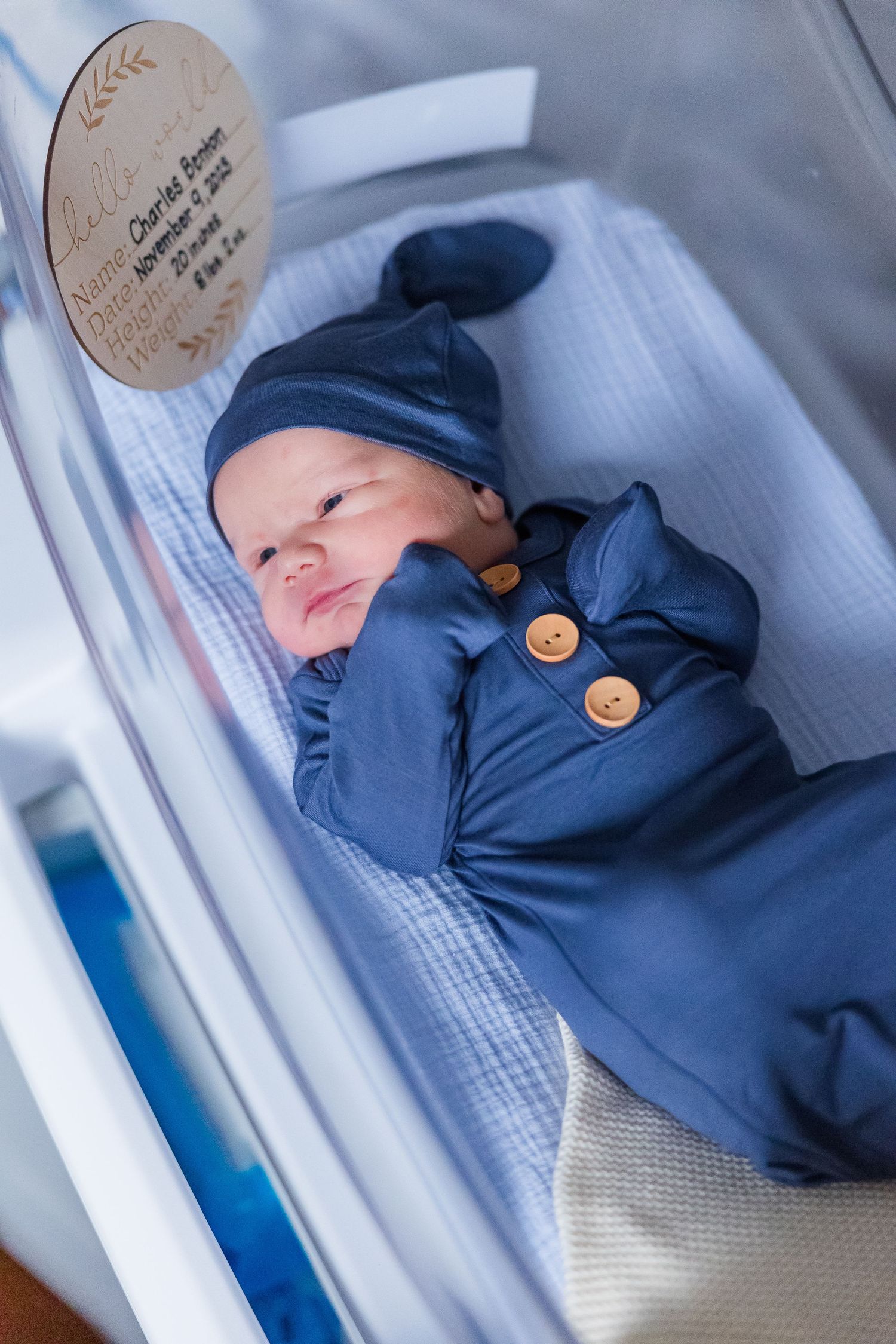 Newborn baby sleeping peacefully in blue outfit with decorative wooden tag.