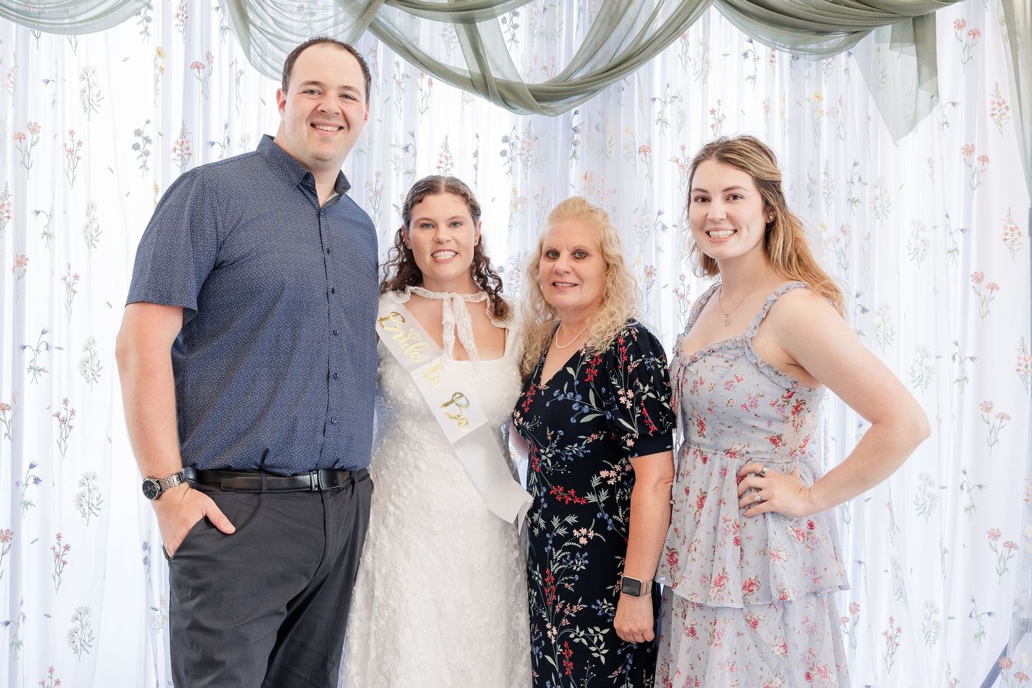 Bride in white dress smiling with three family members in front of sheer curtain wedding backdrop.