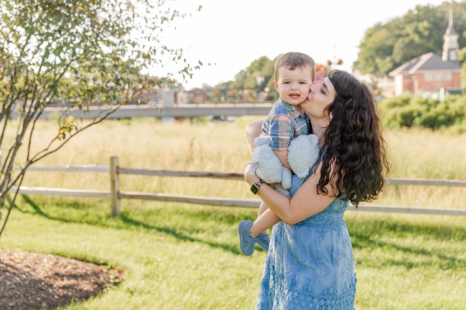 A mother in a blue dress kisses her smiling baby boy outdoors near a wooden fence on a sunny day.