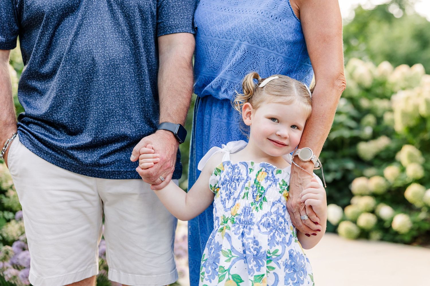 Toddler girl in floral dress holds hands with parents near hydrangea bushes.