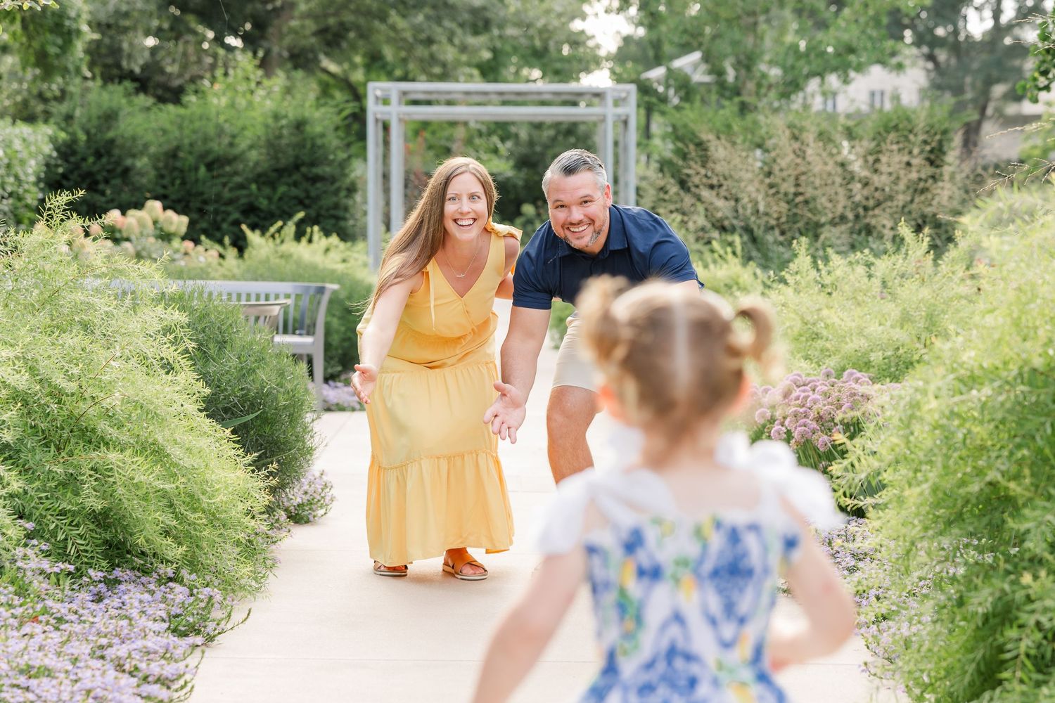 A joyful family in a garden, parents in yellow and navy reaching toward their young daughter on a sunny path.