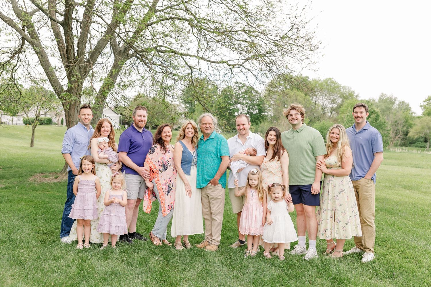 Large multigenerational family posing together outdoors on green grass with trees in background during spring.