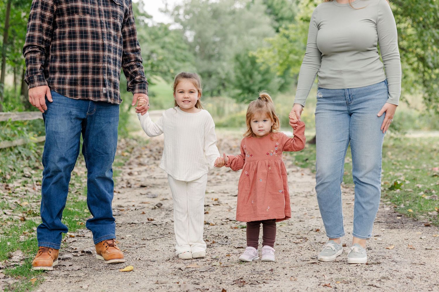 Family of four holding hands on a dirt path in a green park, with two young girls in white and pink outfits.