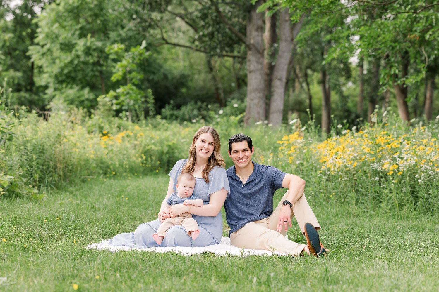 A smiling couple relaxes on a blanket in a green meadow with wildflowers, holding their baby.
