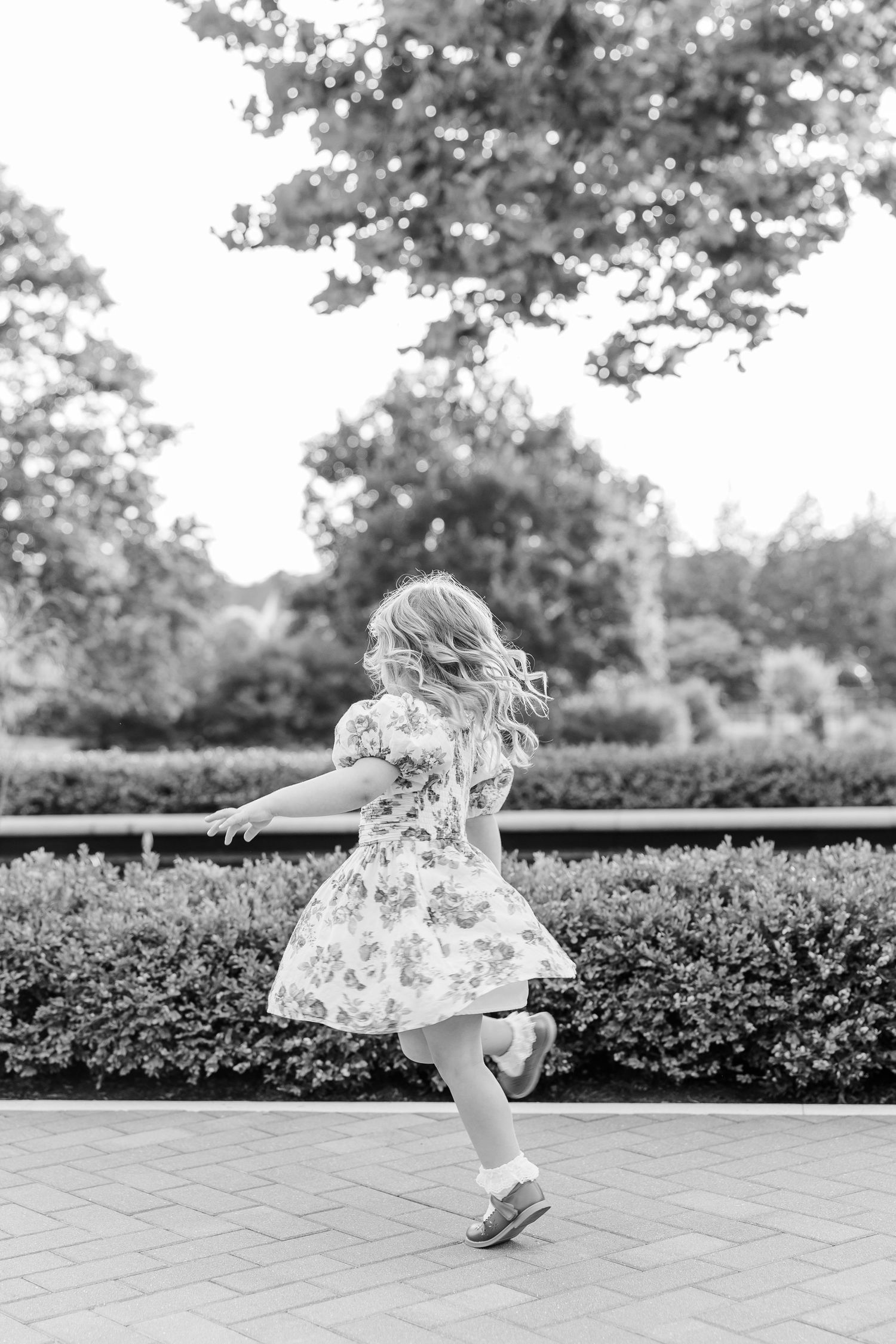 A young girl in a floral dress twirls joyfully on a brick path surrounded by lush garden hedges.
