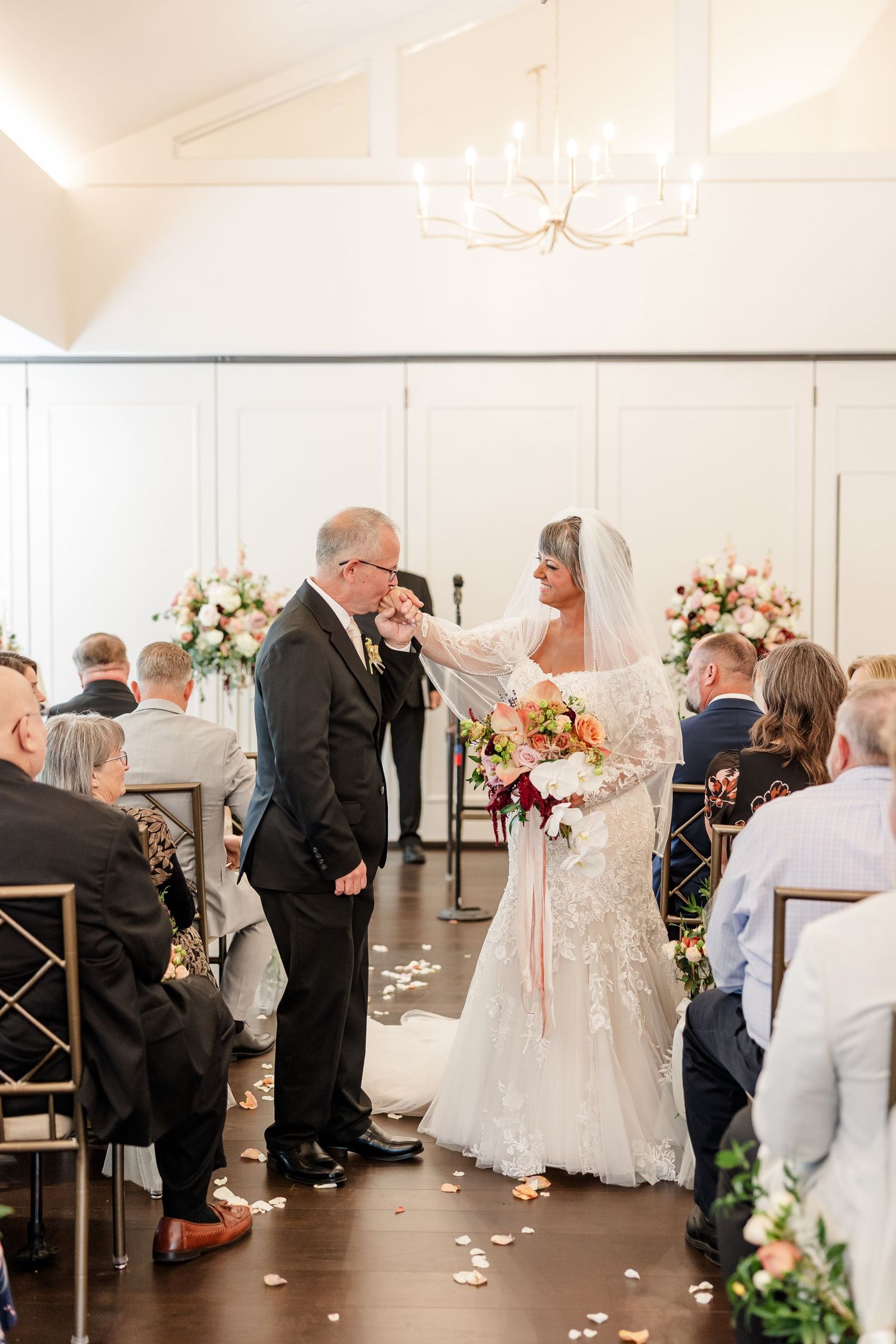 A bride in a lace gown holding a colorful bouquet joins hands with her groom in a dark suit during a wedding ceremony as guests watch.